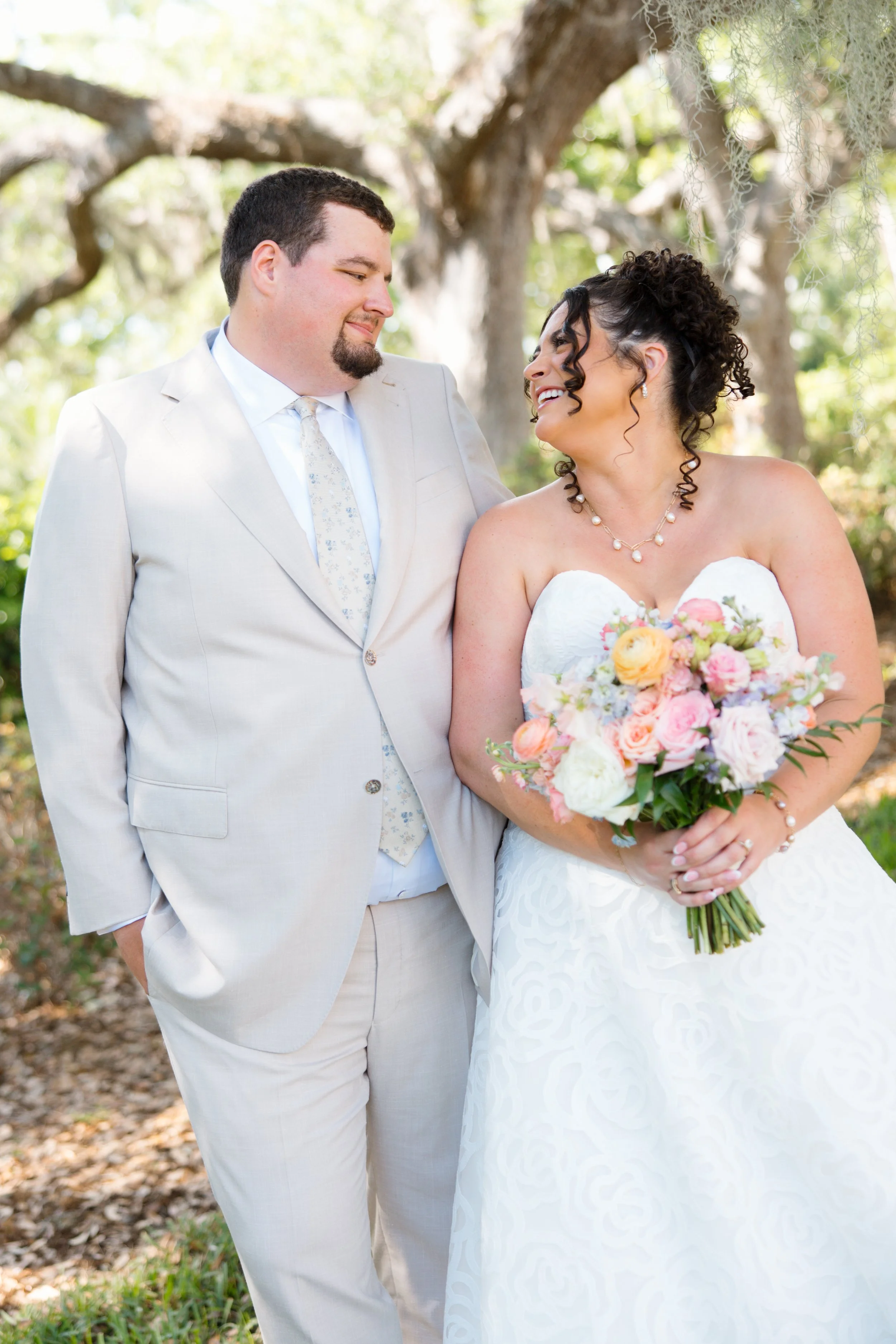 A bride and groom smiling and looking at each other outdoors during their wedding, with trees in the background. The bride is holding a large bouquet of pink, yellow, and white flowers, and wearing a white strapless wedding dress with a pearl necklac