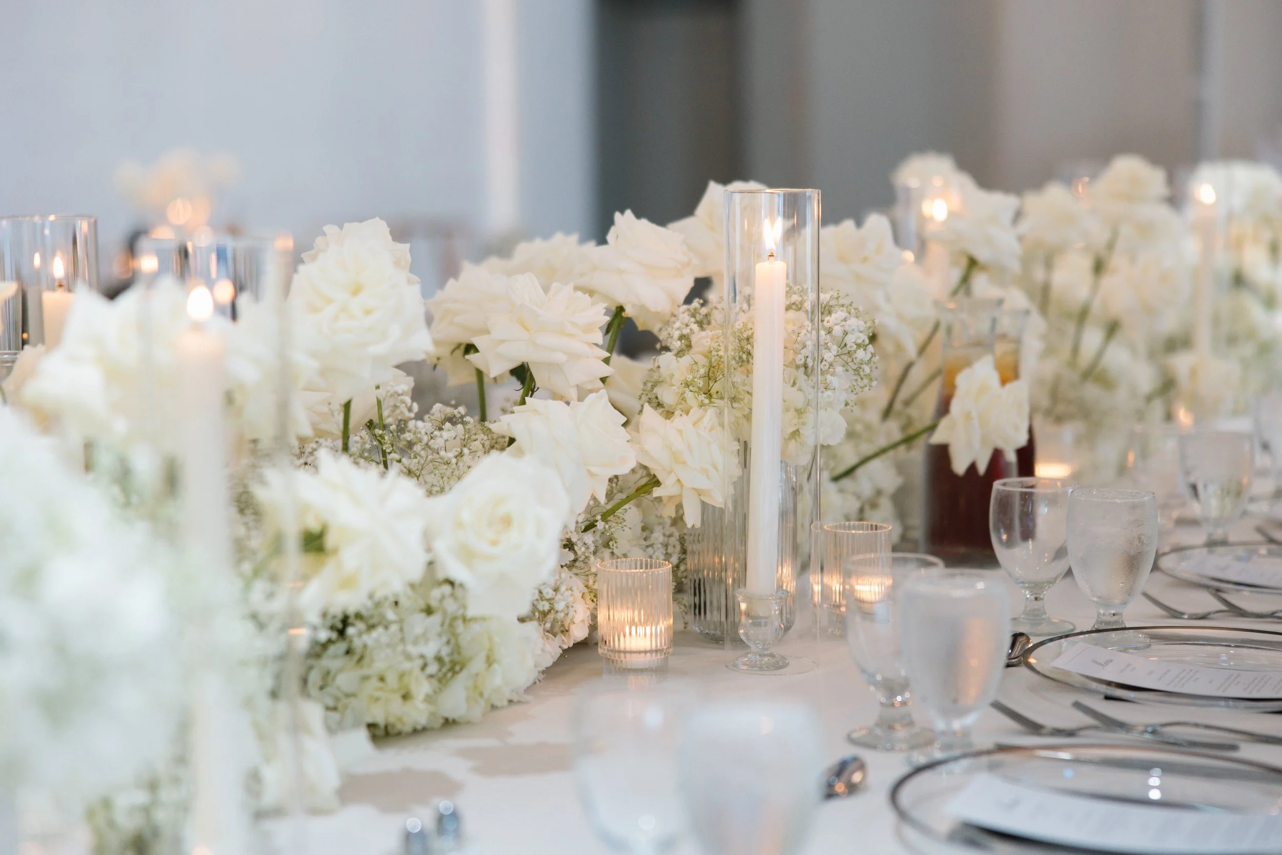 A table decorated with white flowers, tea light candles in glass holders, and tall cylindrical candles in glass containers, set for a formal event or wedding.