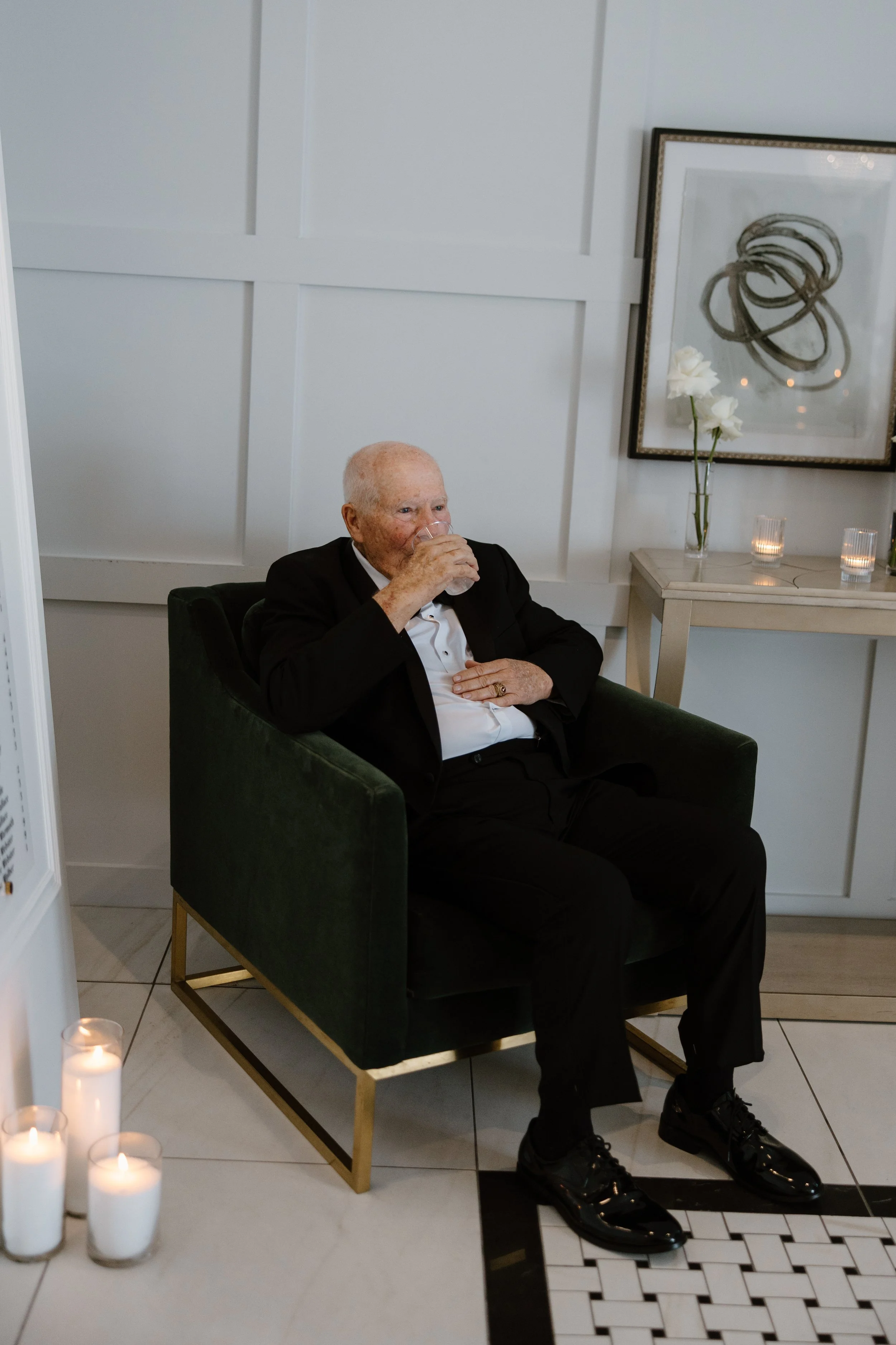 An elderly man in a tuxedo sitting in a green armchair, drinking from a glass. The setting appears to be a decorated indoor space with a table that has white roses, candles, and framed artwork on the wall.