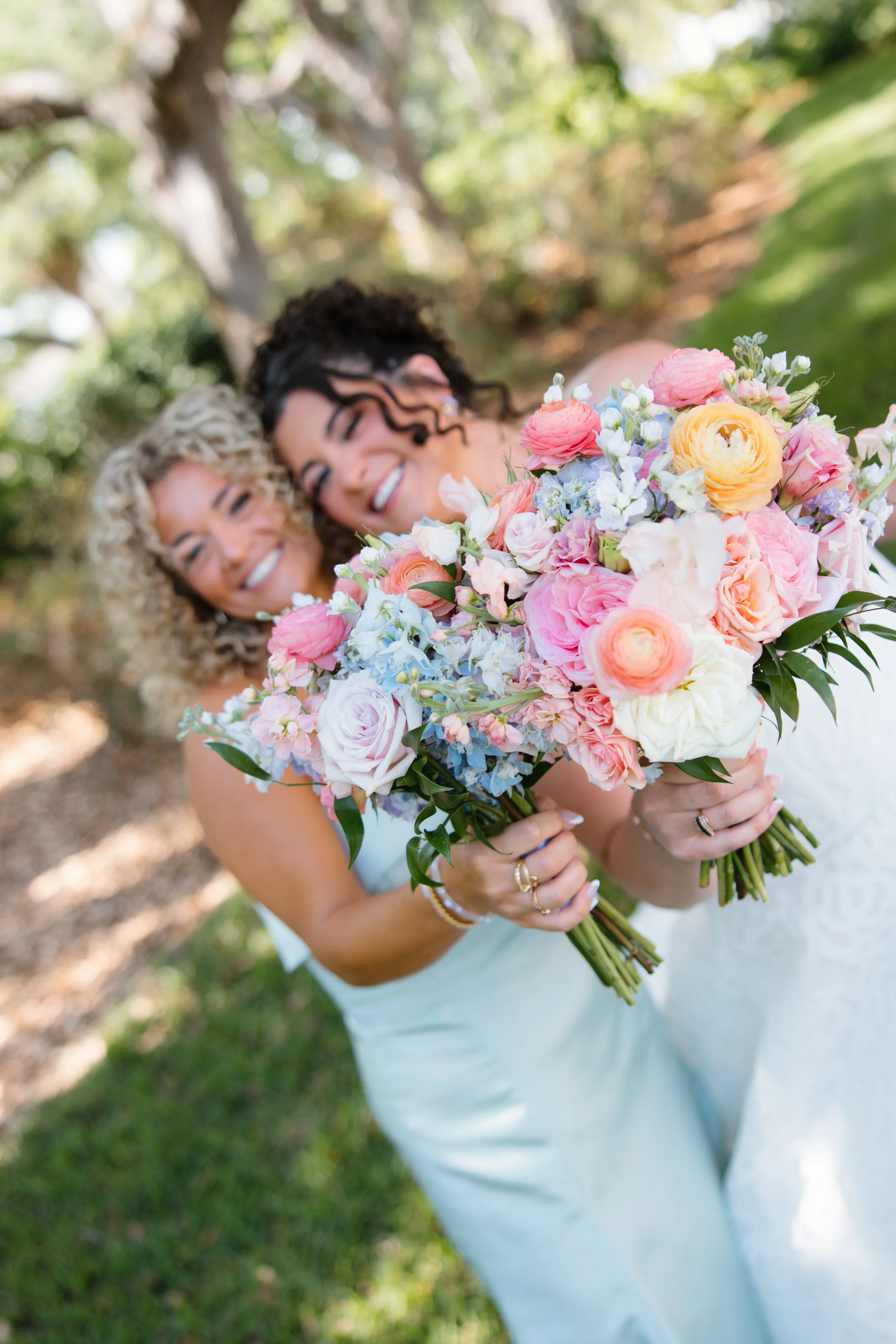 Two smiling women holding a large bouquet of pink, peach, white, and blue flowers outside on a sunny day.