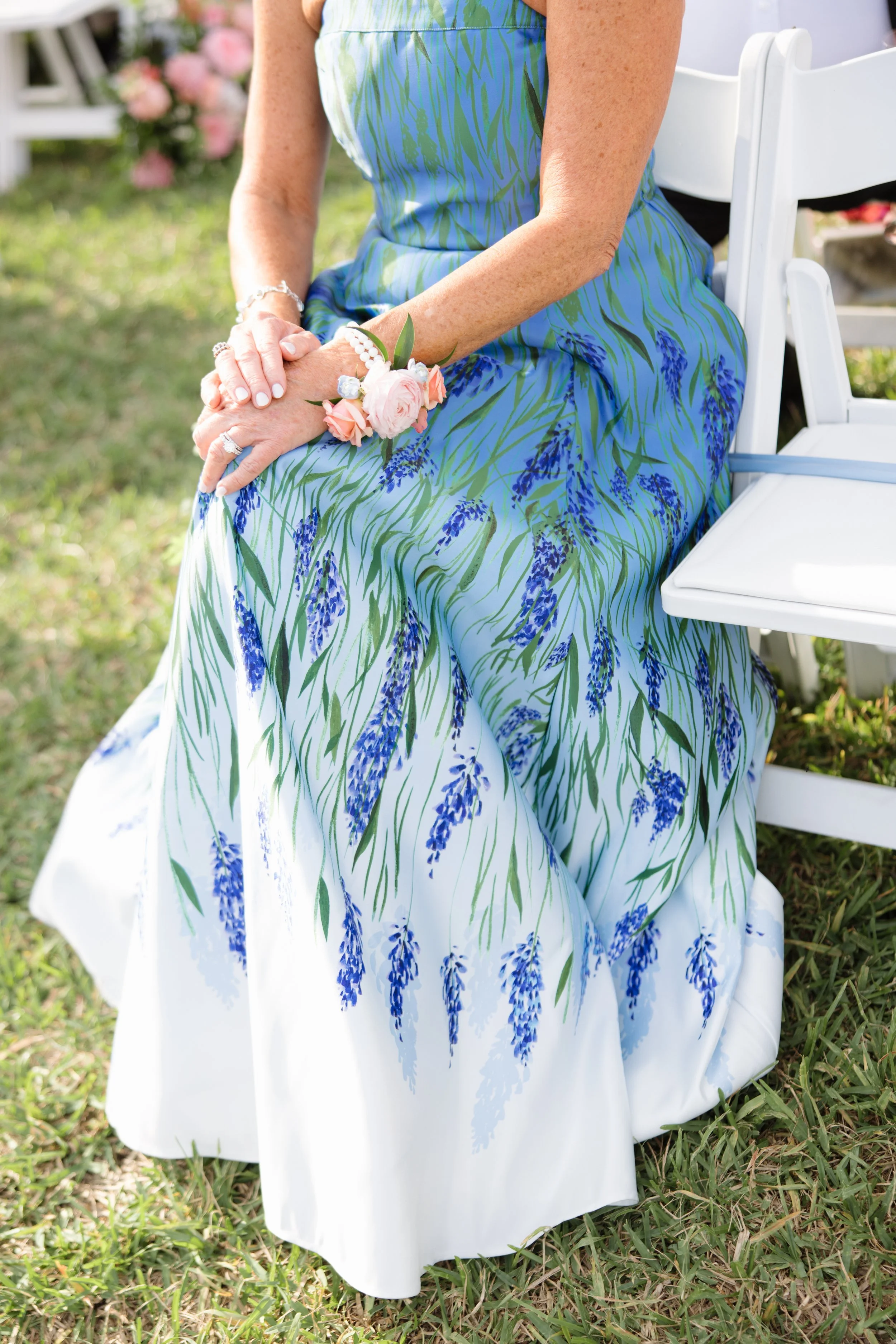 Close-up of a woman in a floral blue and green dress sitting on a white chair outdoors, with her hands clasped and wearing a floral wrist corsage.