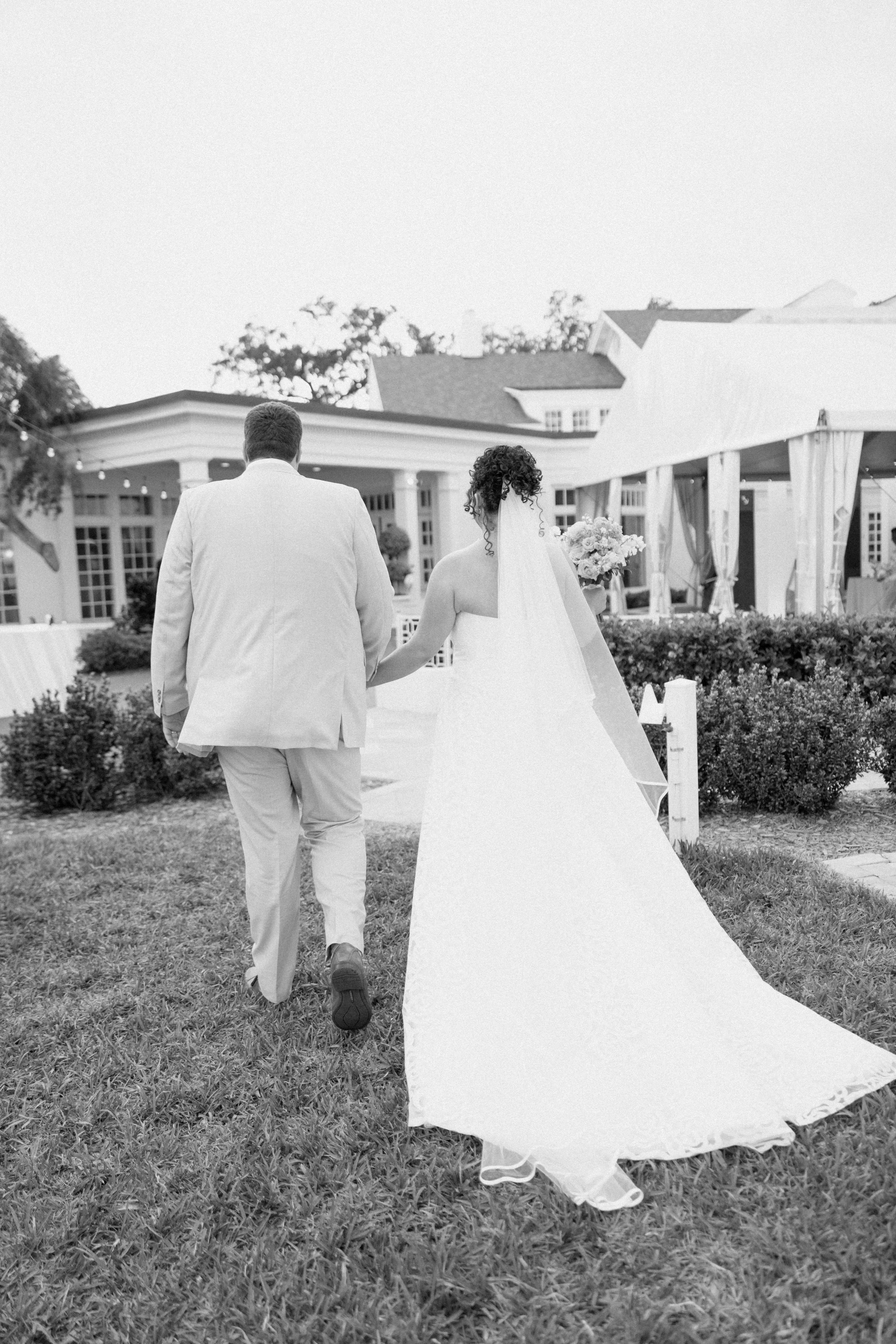 A bride and groom walking hand in hand outdoors near a wedding reception area with a tent and building in the background.