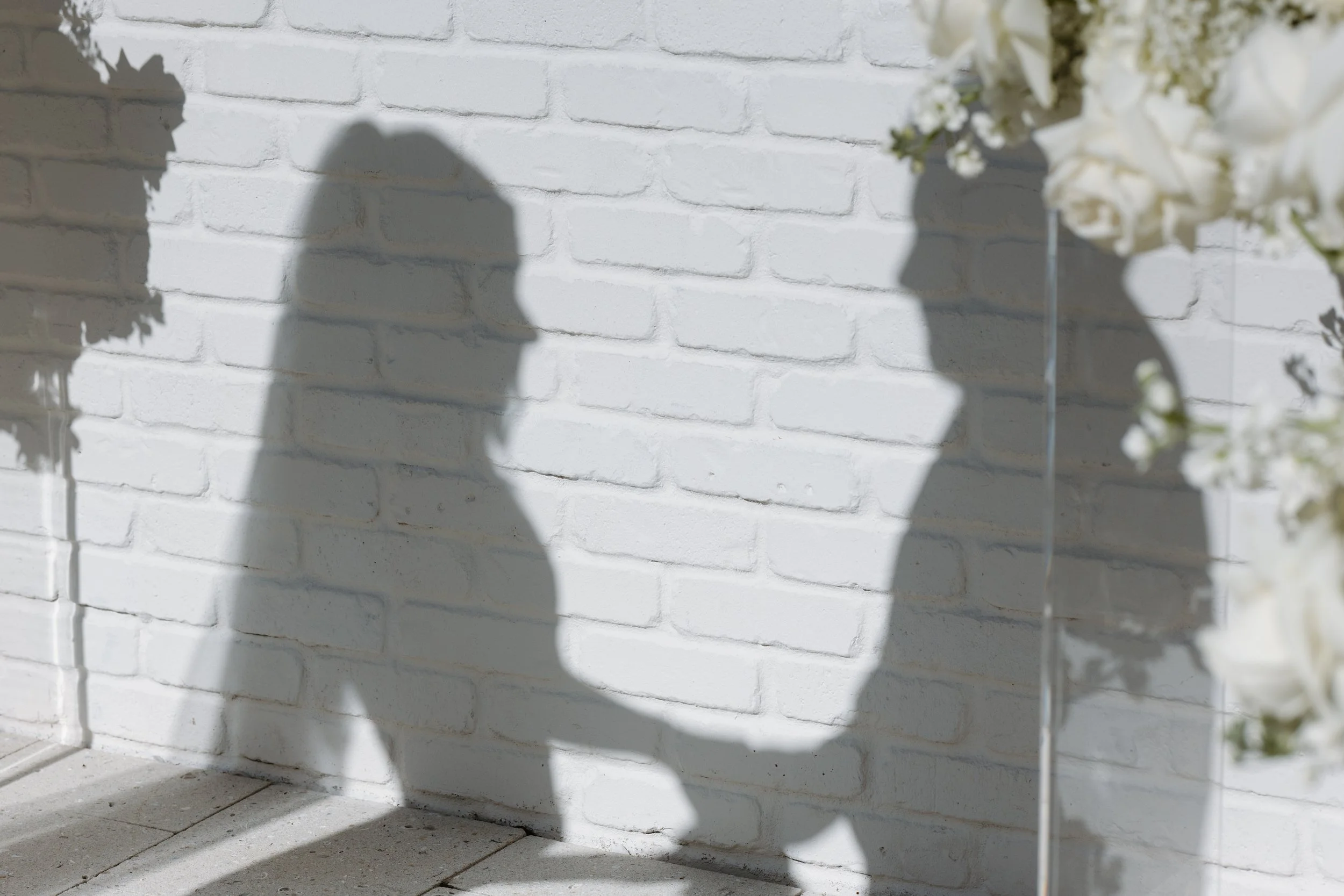 Shadows of a person and floral arrangements cast on a white brick wall illuminated by sunlight.