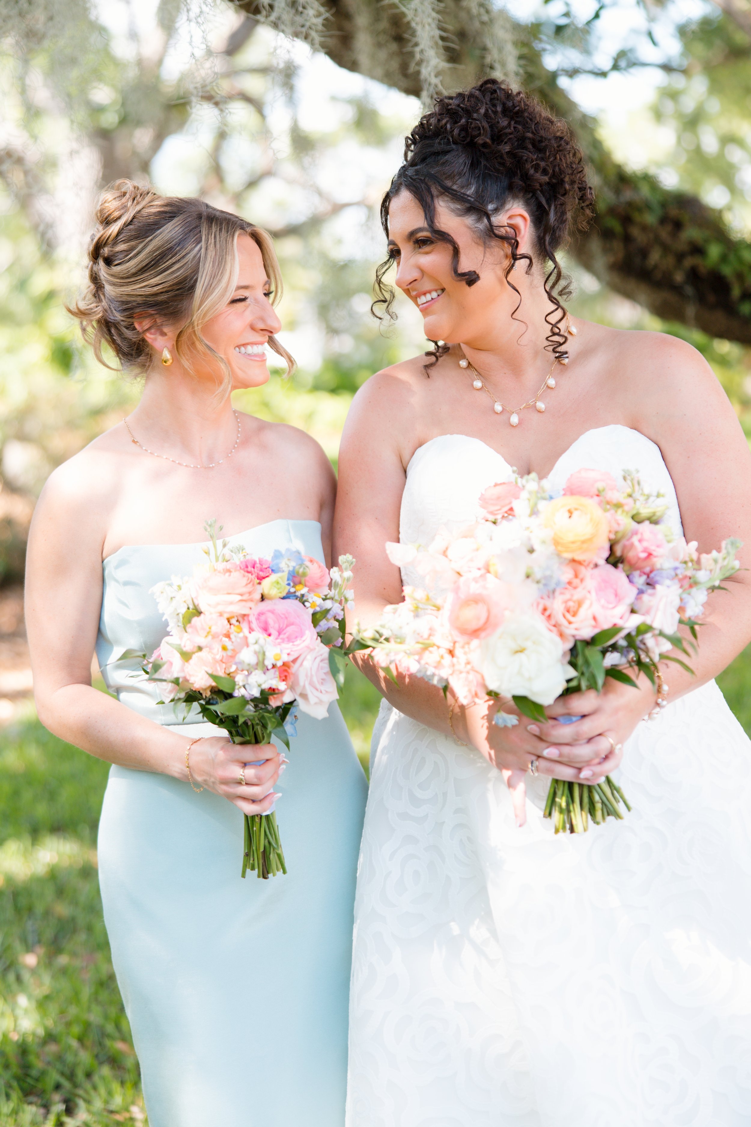 Two women in wedding dresses smiling and holding bouquets of pink, white, and pastel-colored flowers outdoors with green trees in the background.