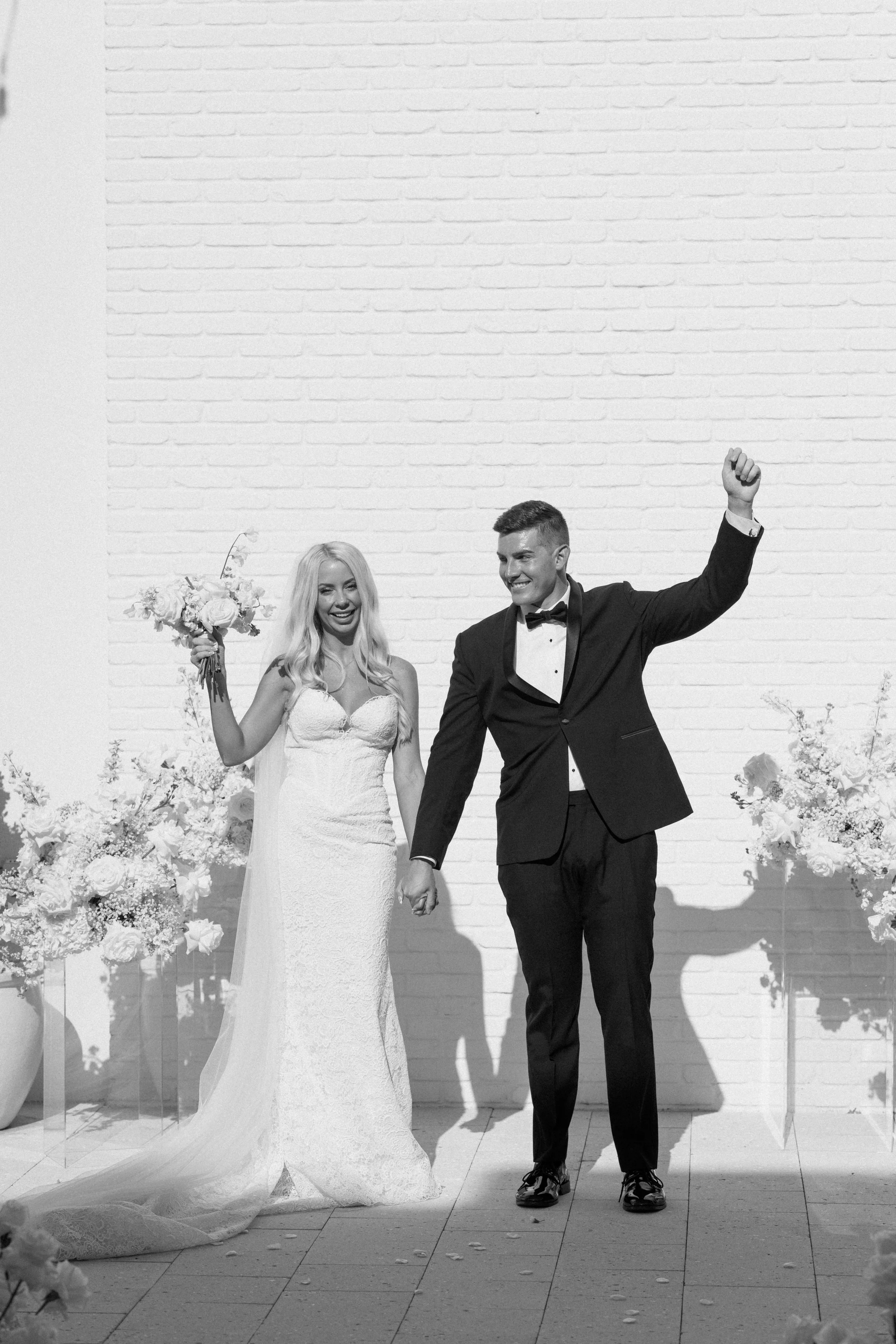 A bride and groom holding hands and smiling at their wedding ceremony, with flowers and decorative elements around them, against a white brick wall background.