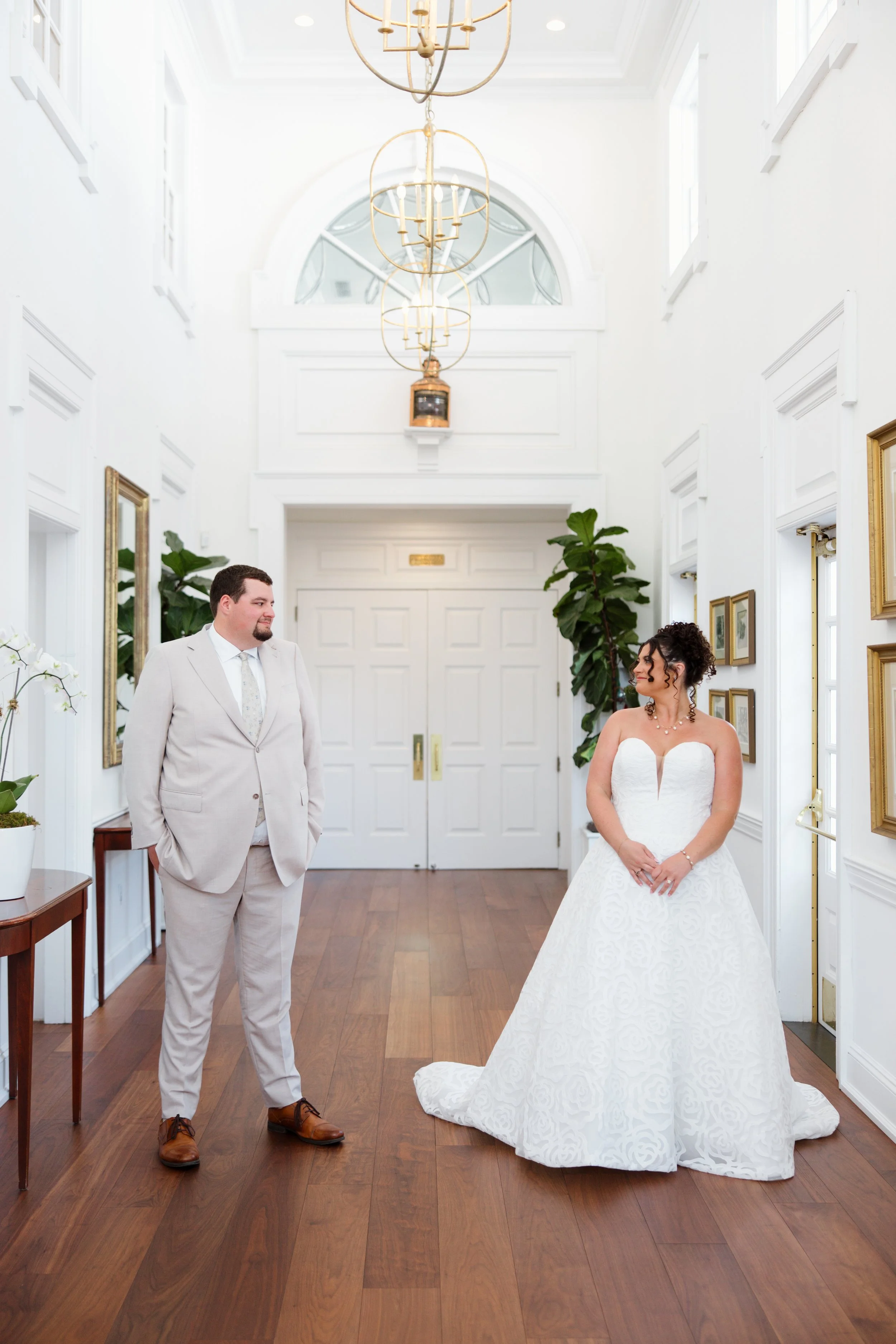 A bride and groom standing in a bright, elegant hallway with white walls, large windows, and wooden flooring, during their wedding ceremony.