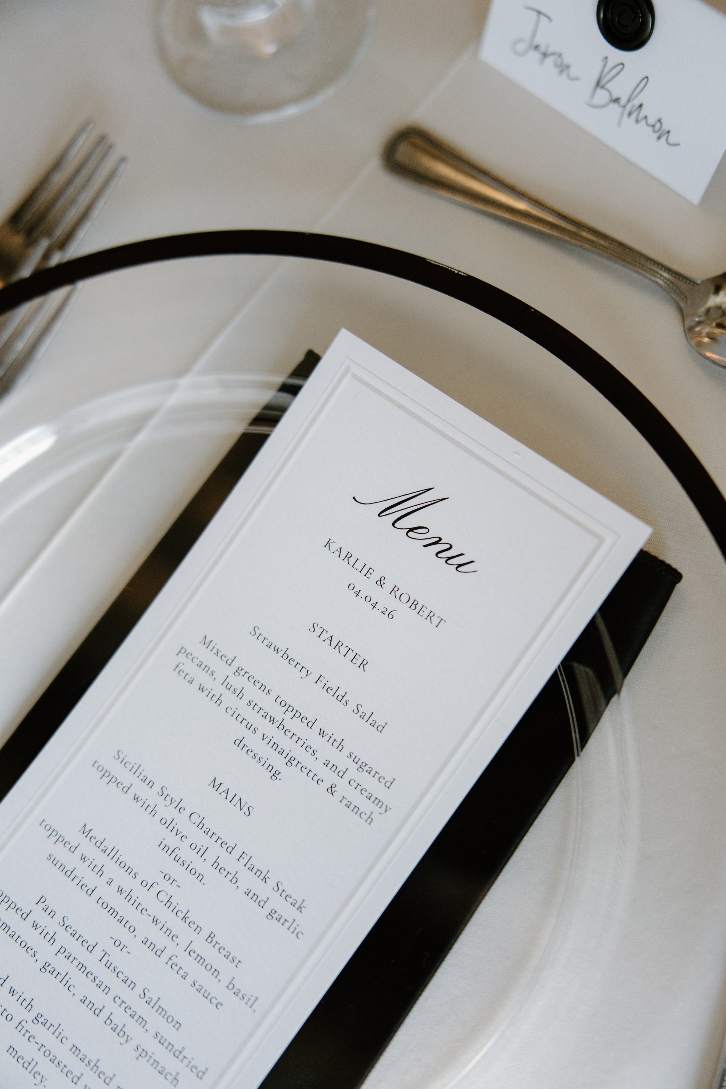 Close-up of a printed menu with a white background, black text, and elegant font, featuring a description of a strawberry Fields Salad and other dishes, placed on a table with silverware and a wine glass nearby.