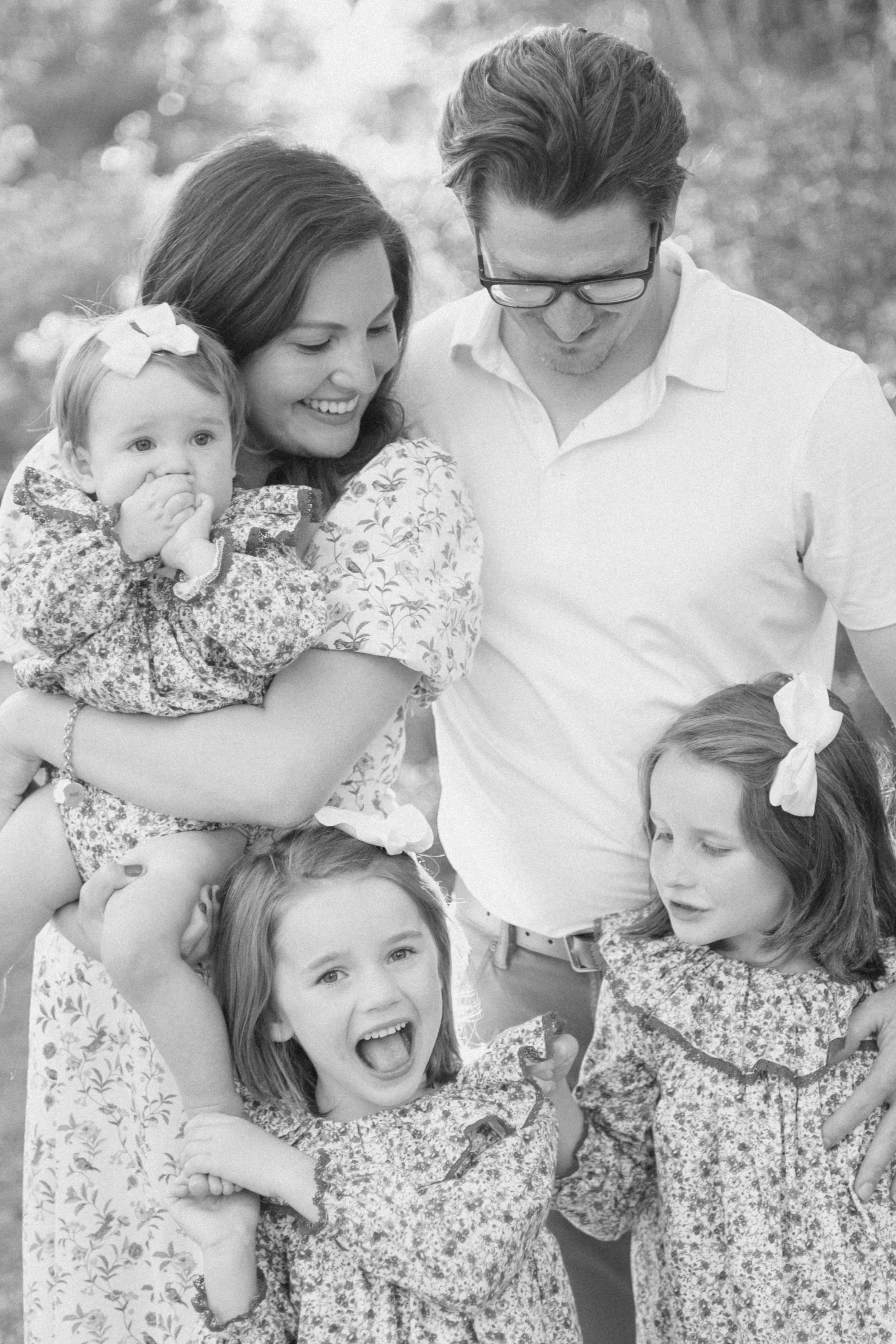 A black and white photo of a family, including a woman holding a young girl with a bow in her hair, a man with glasses, and a girl with a bow in her hair, all smiling and interacting outdoors.
