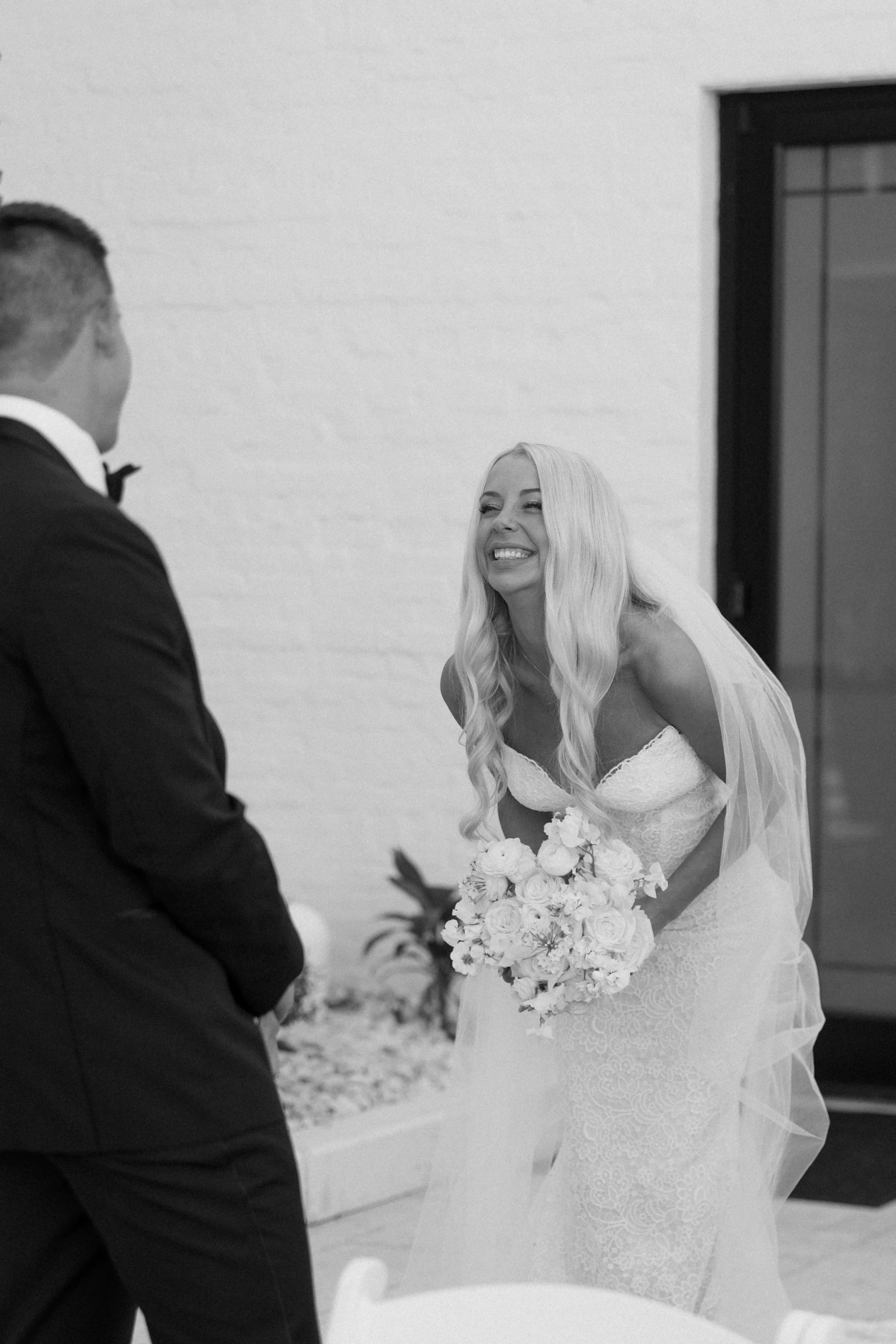 A bride with long blonde hair wearing a strapless lace wedding dress holding a bouquet of white roses, smiling during a wedding ceremony.