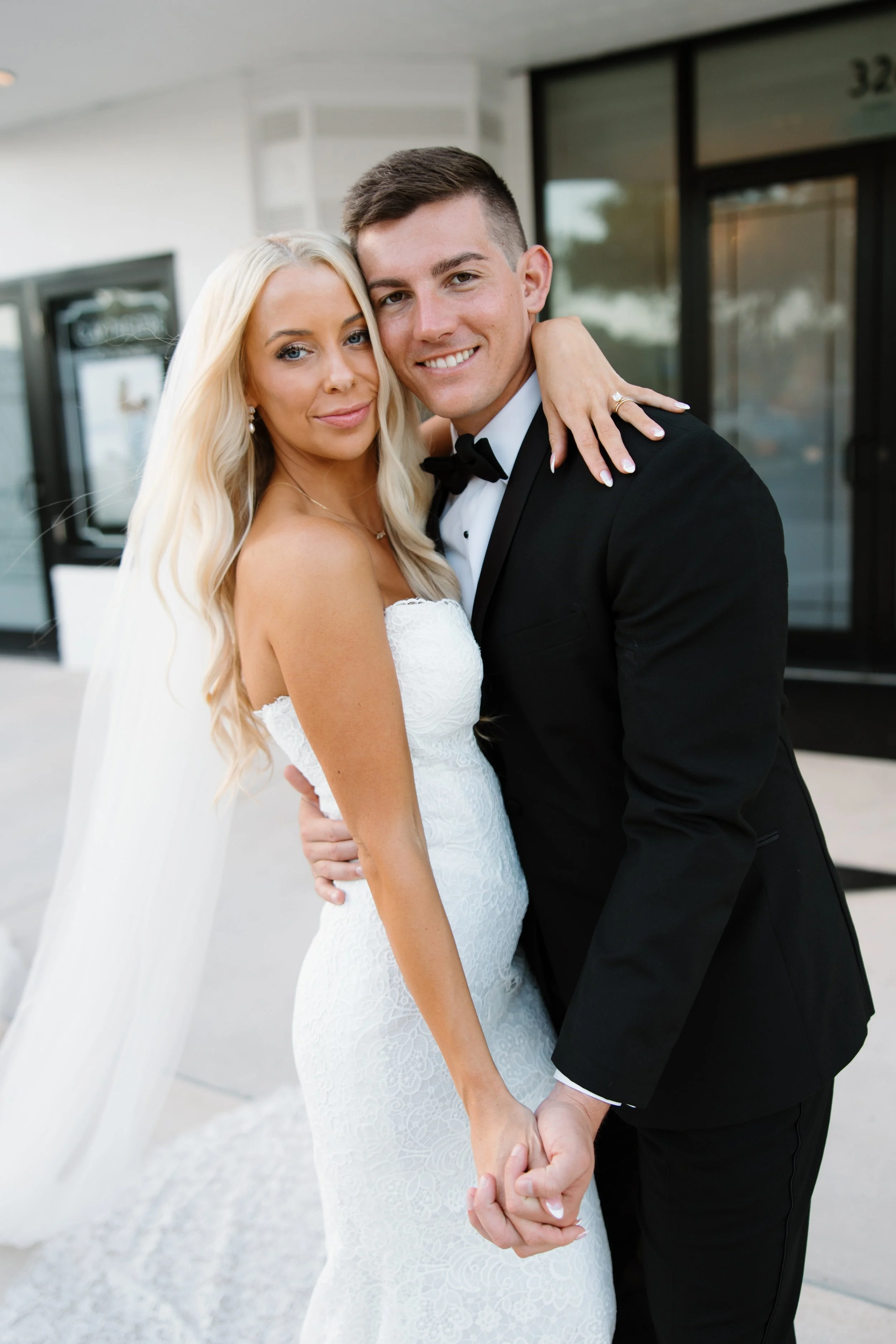 A bride and groom in wedding attire smiling and holding hands outside a building with glass doors.
