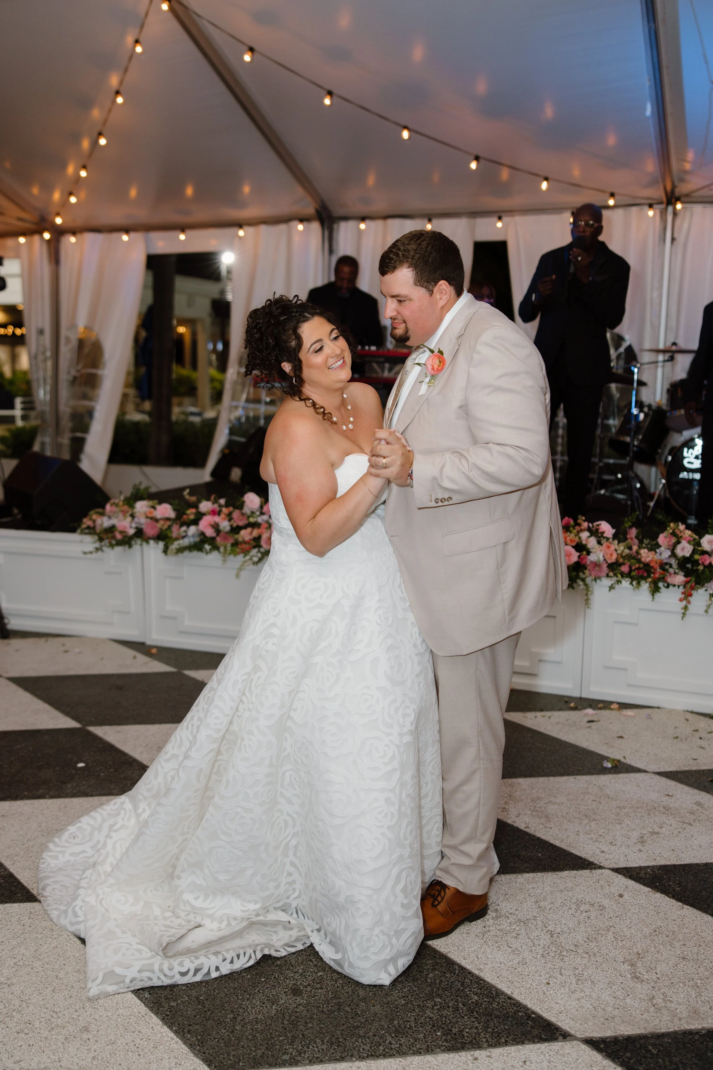 A wedding couple dancing under a tent with string lights, with a decorated table with flowers behind them.