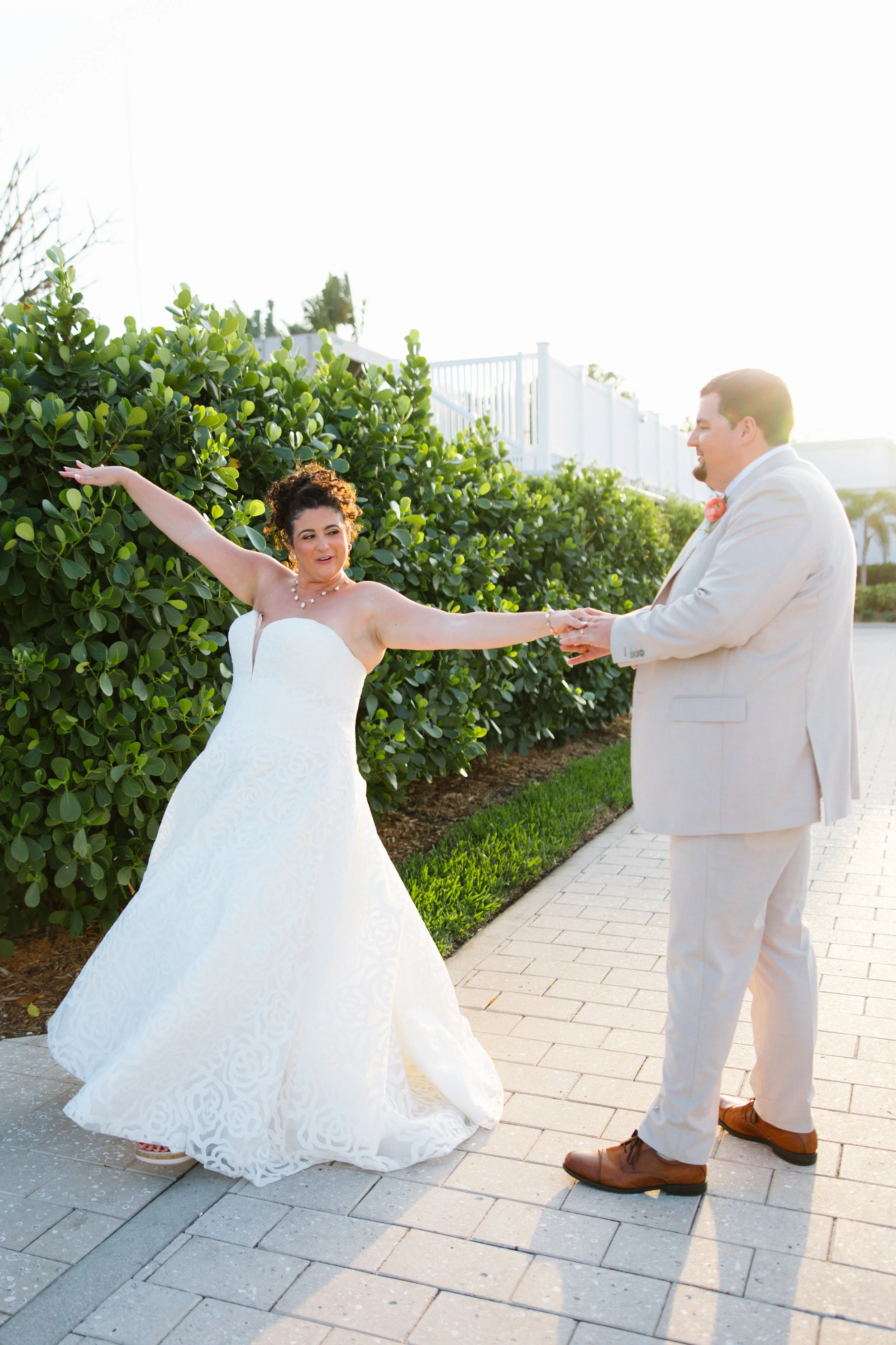 A bride and groom holding hands outdoors, with the bride raising one arm and the groom smiling. The bride is wearing a strapless white wedding gown, and the groom is in a light-colored suit with a pink boutonniere. Bright sunlight creates a glow arou