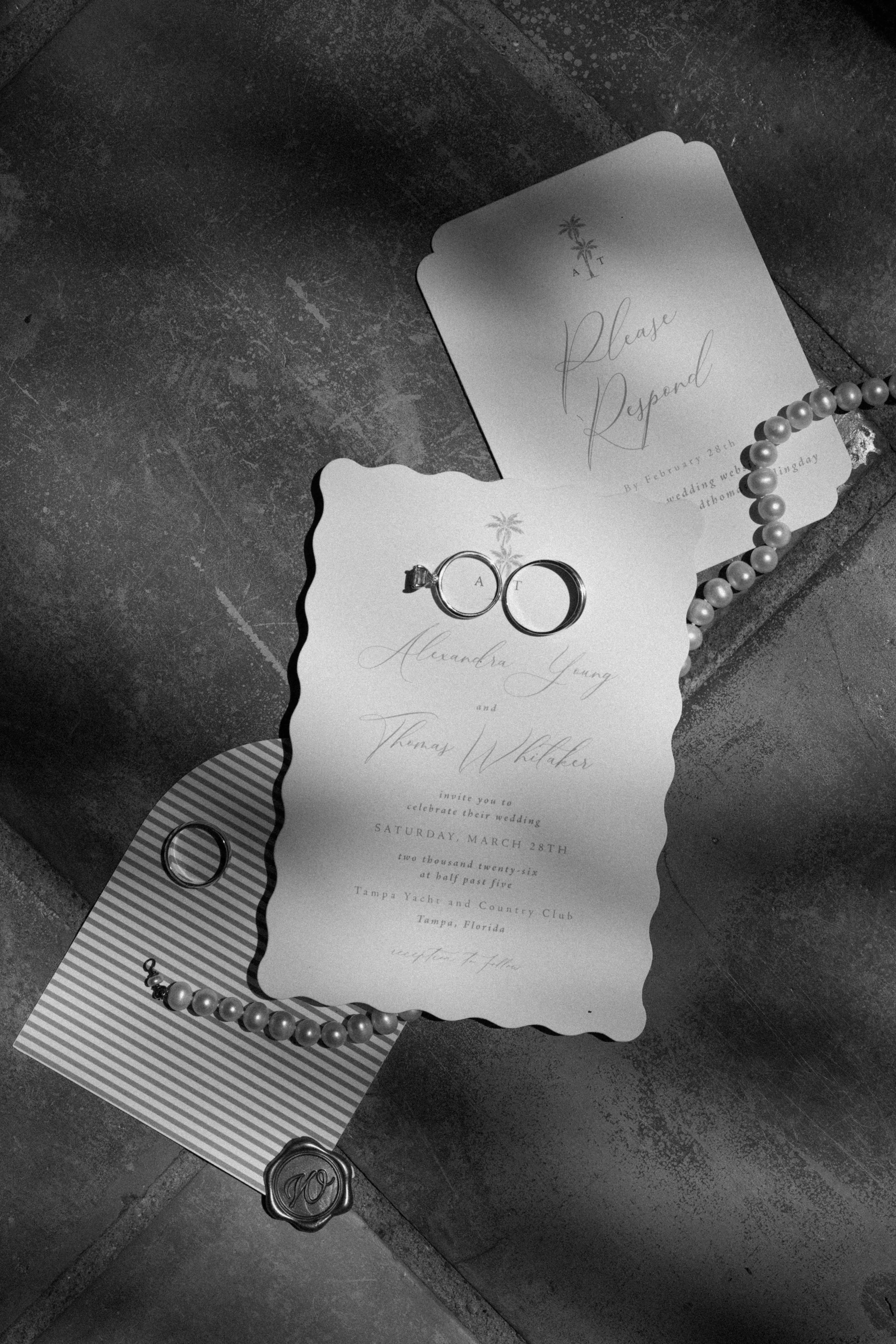 Wedding invitation card with a pearl necklace, wedding rings, and a wax seal on a grey tiled surface.