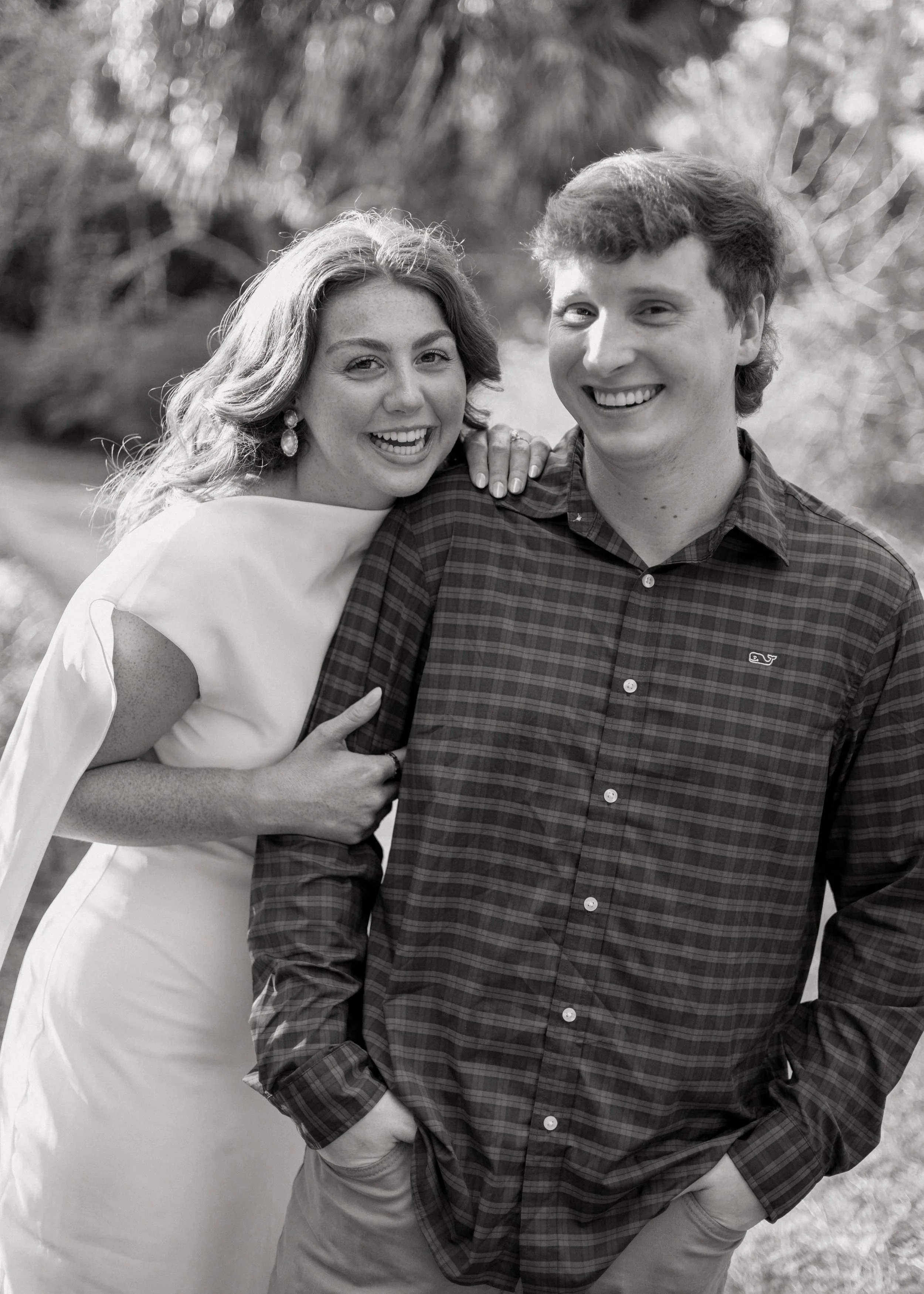 A smiling young woman with wavy hair and earrings leaning on a young man with short hair, wearing a plaid shirt. They are outdoors with blurred trees in the background.