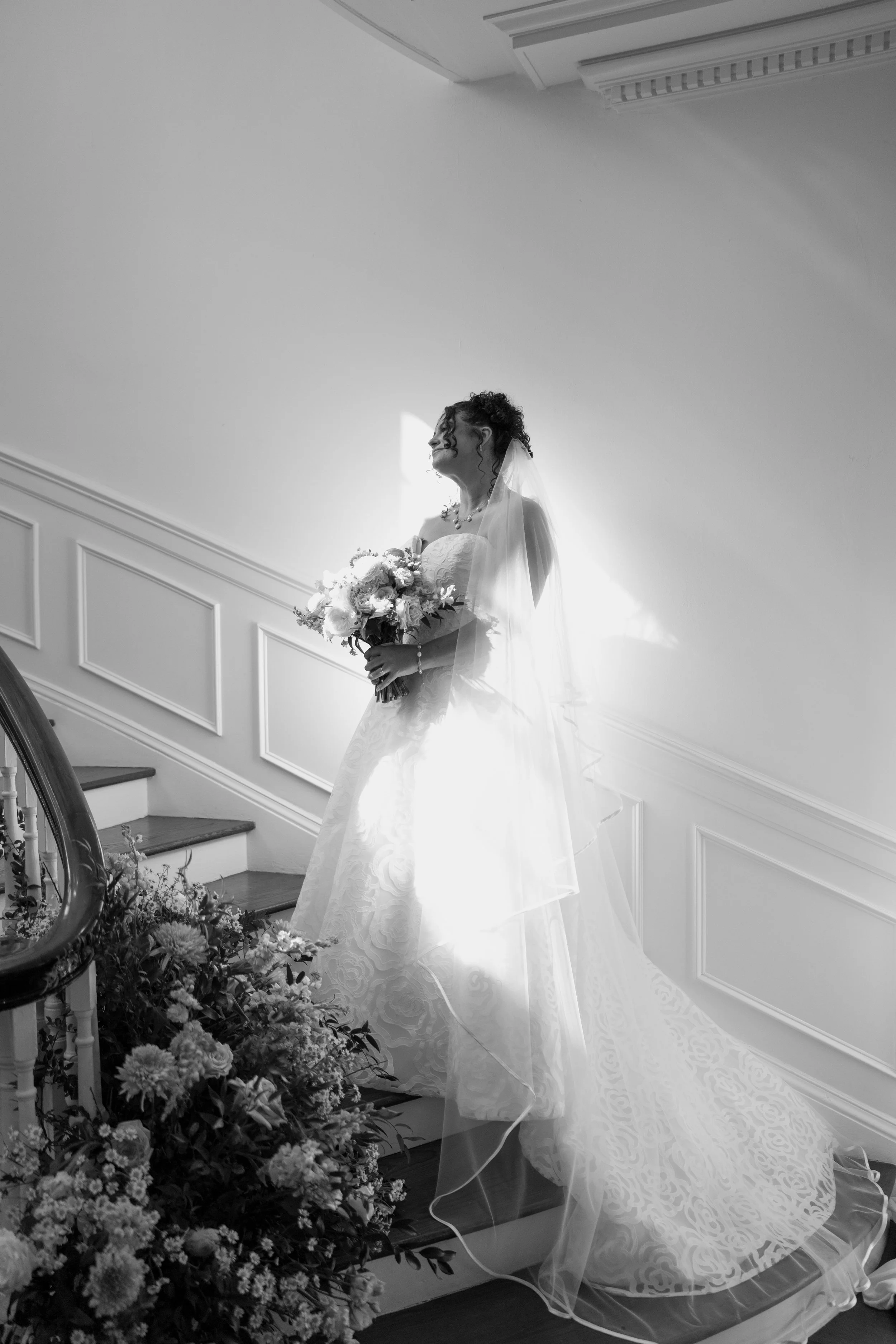 A bride standing on a staircase holding a bouquet of flowers, wearing a wedding dress with a veil, surrounded by floral arrangements, with sunlight shining behind her.
