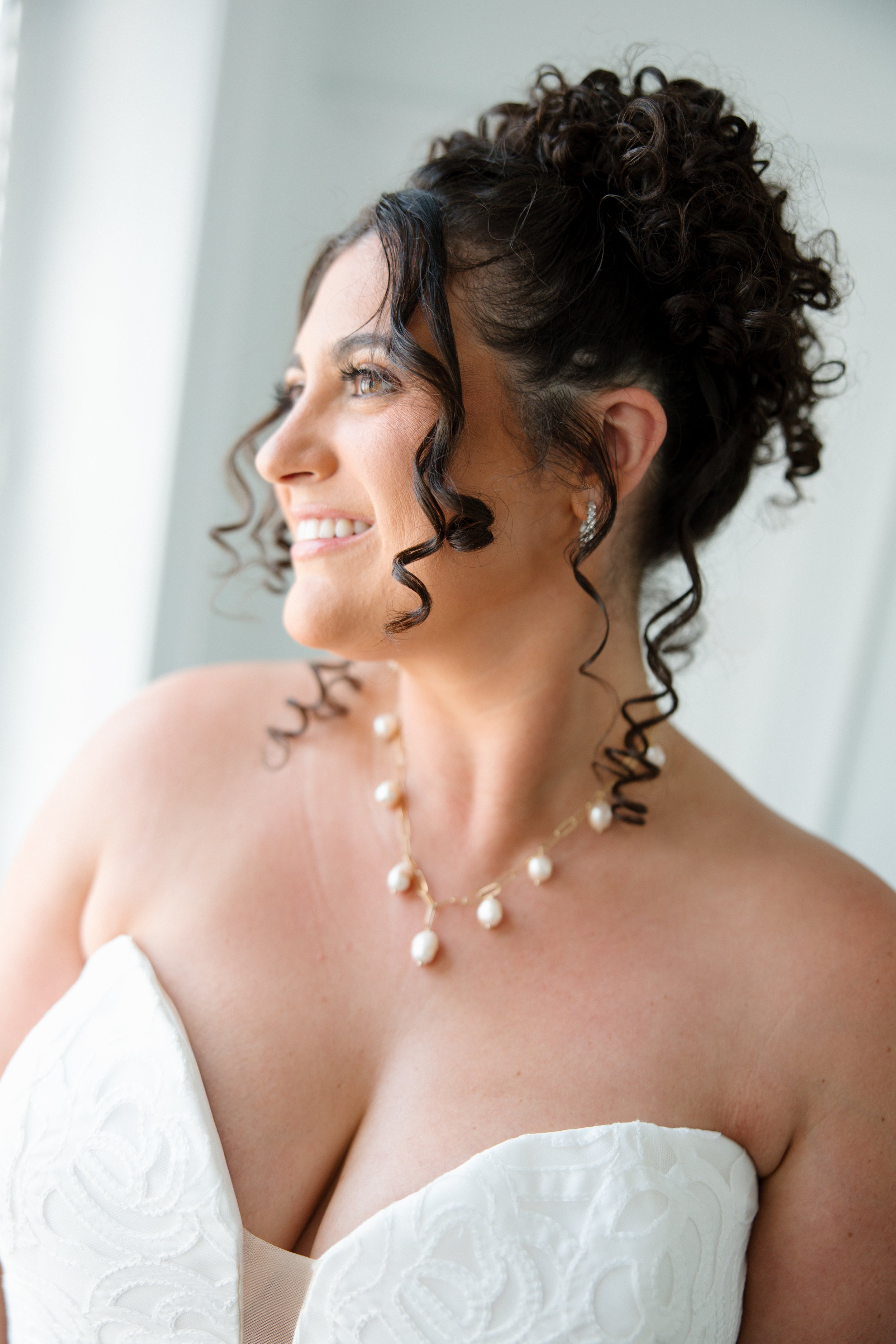 Close-up of a woman with curly brown hair and blue eyes, wearing pearl jewelry and a white strapless dress, looking to the left and smiling.