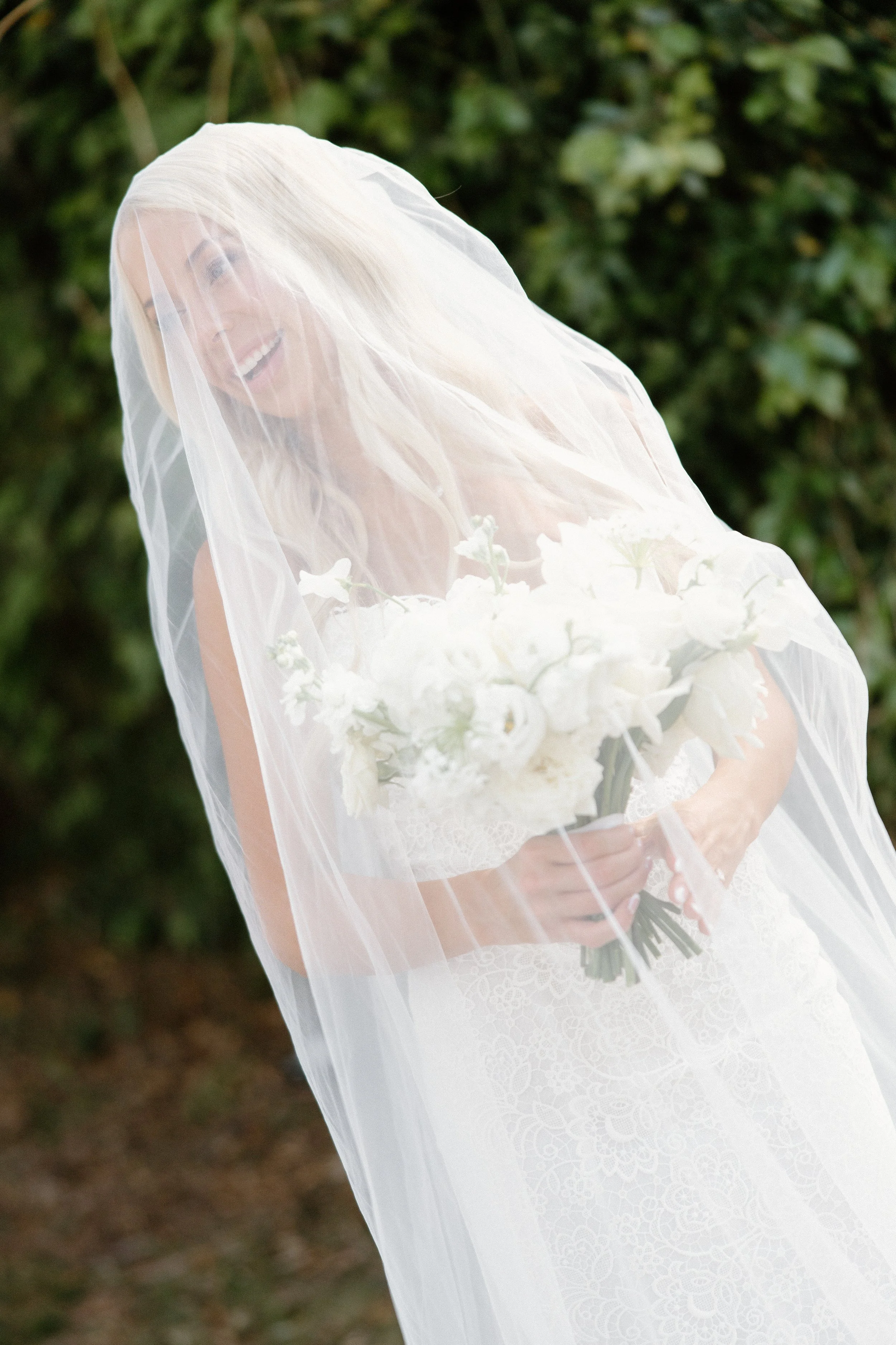 A smiling woman on her wedding day, wearing a white lace wedding dress and veil, holding a bouquet of white flowers, standing outdoors with blurred green foliage in the background.