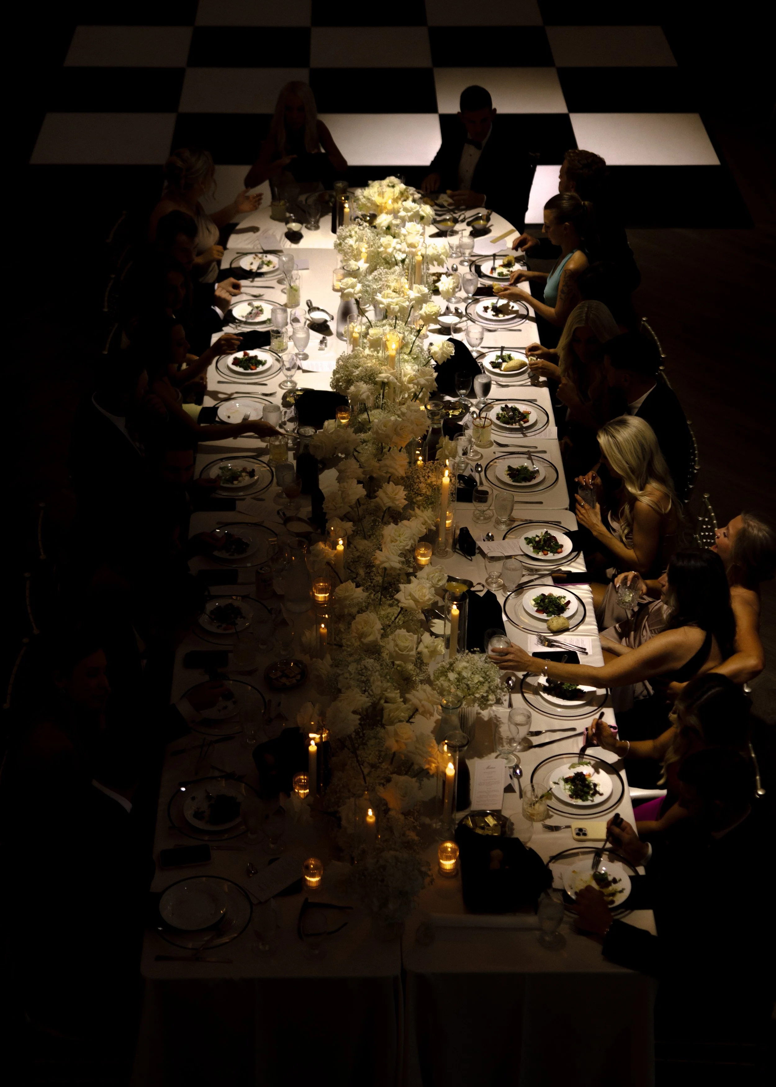 A formal dinner party with people sitting around a long table decorated with white flowers and candlelit lanterns, with a black and white checkered floor.