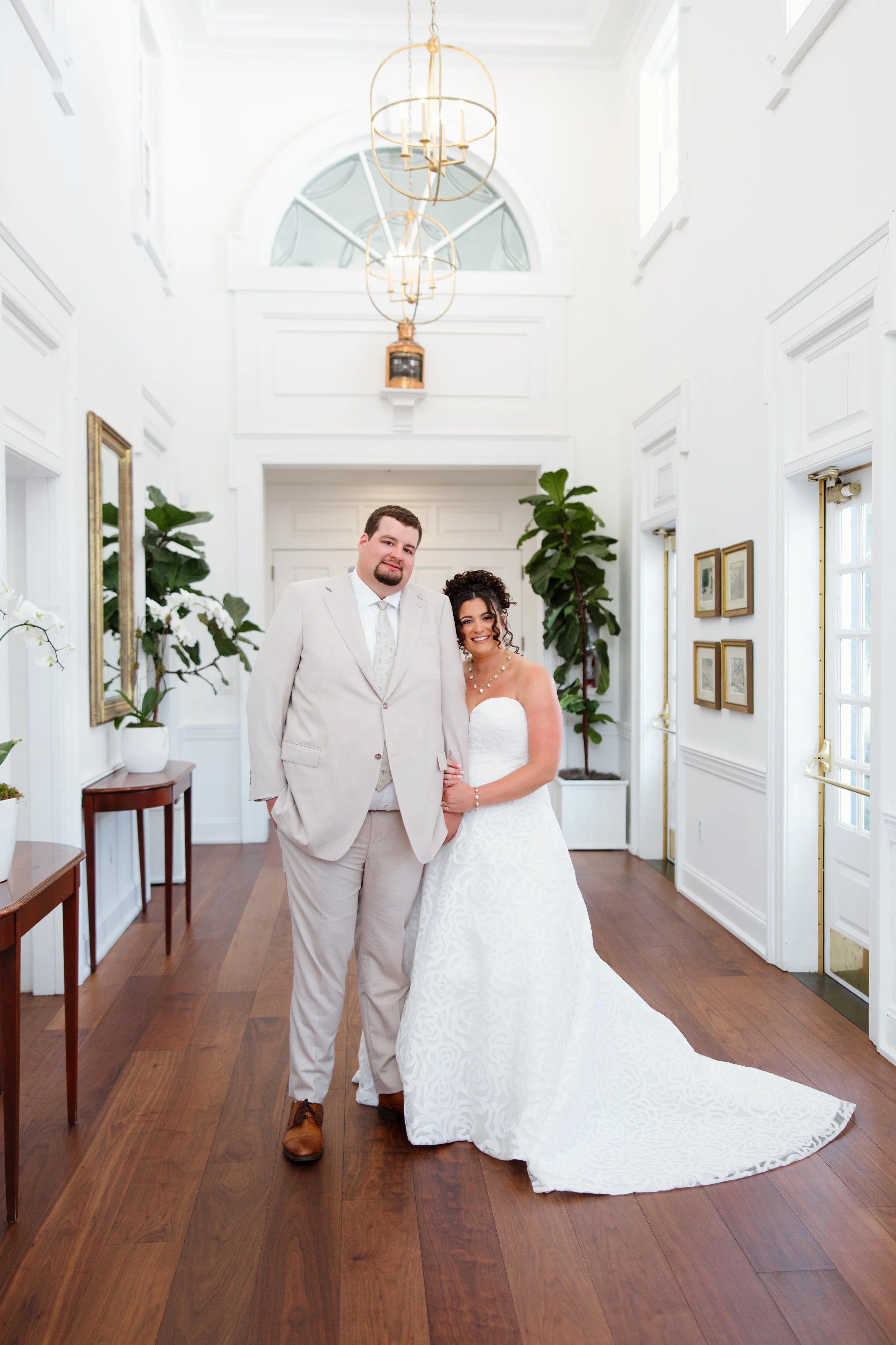 A couple dressed in wedding attire standing inside a bright, white-walled room with tall windows and wooden floors, flanked by potted plants and framed artwork.