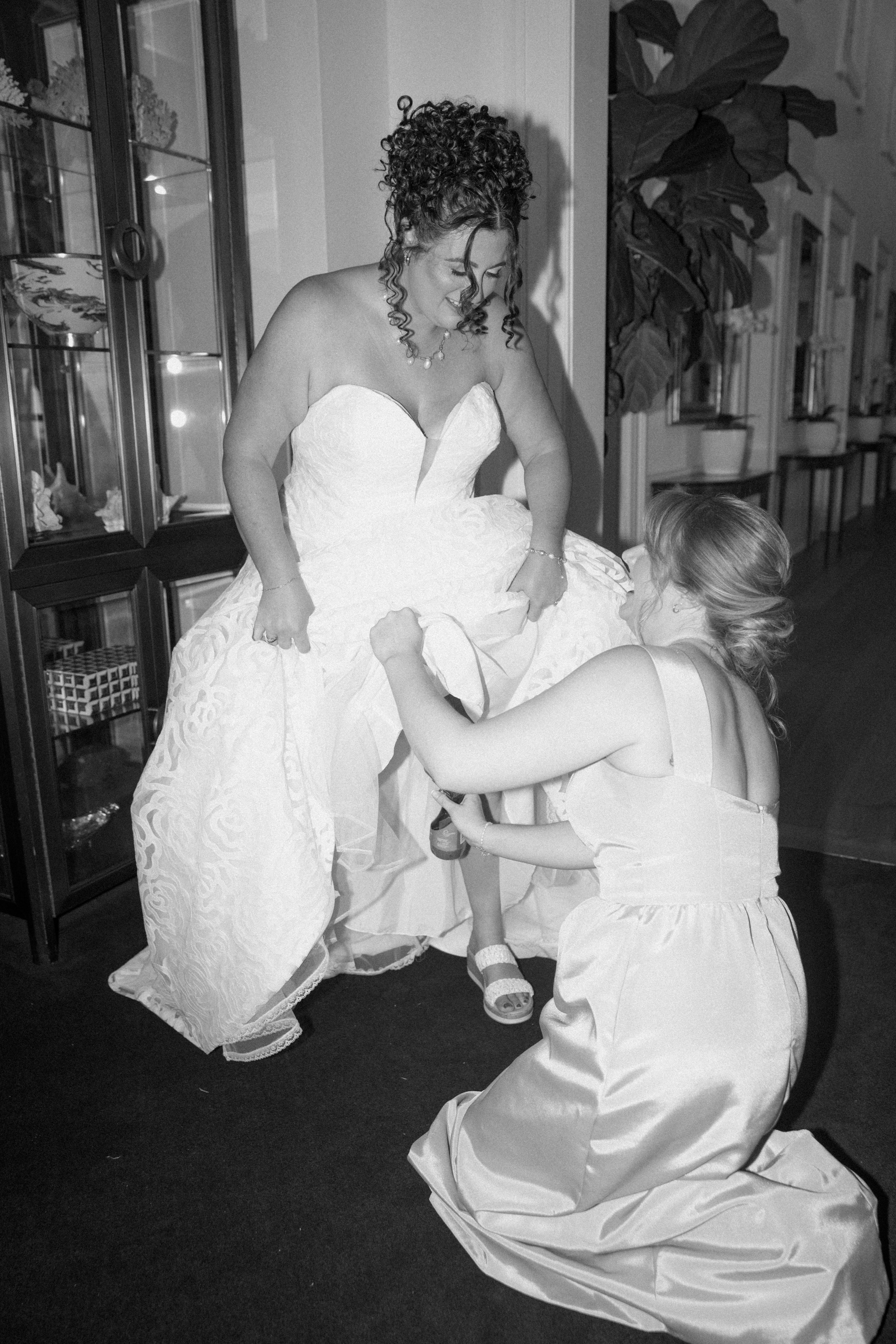 A woman in a wedding dress being helped with her gown by another woman in a strapless dress, inside a house. The bride has curly hair styled in an updo, and the other woman has her hair in a loose style. They are smiling and appear to be enjoying the