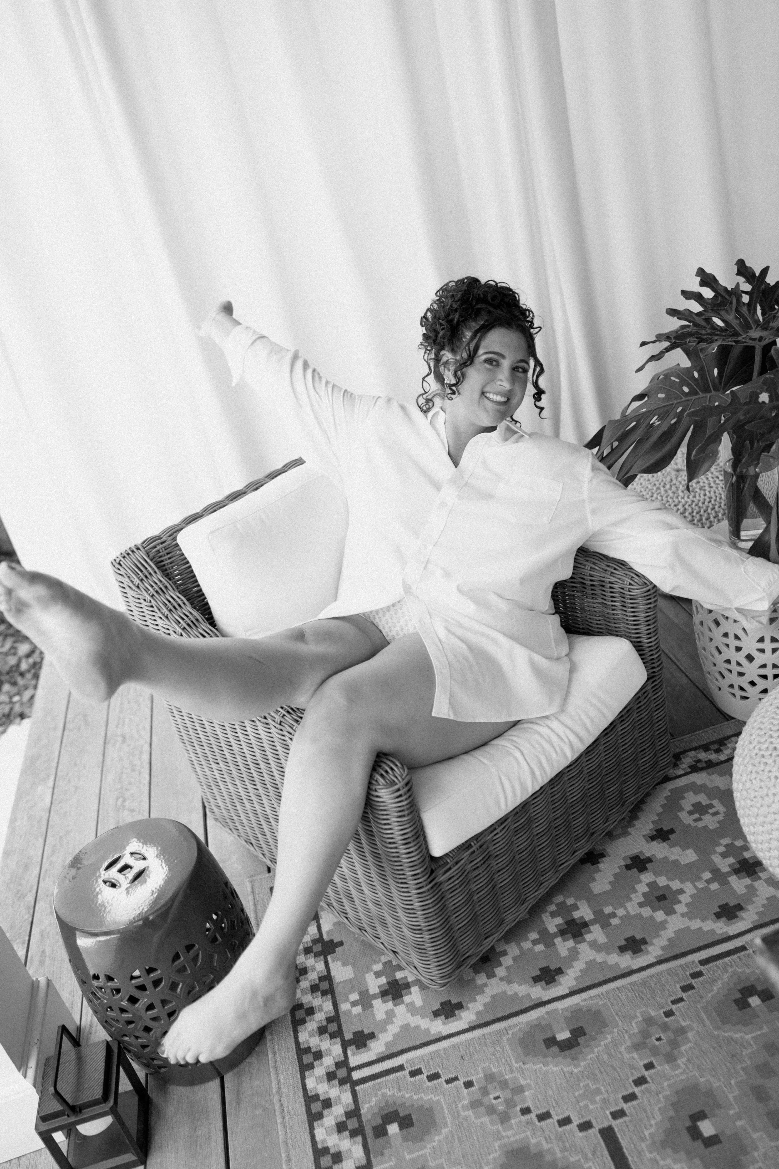 A smiling woman with curly hair sitting on a wicker sofa, wearing a white oversized shirt and shorts, stretching her arms and legs out, in a well-lit room with a patterned rug, tall curtains, and a large plant.