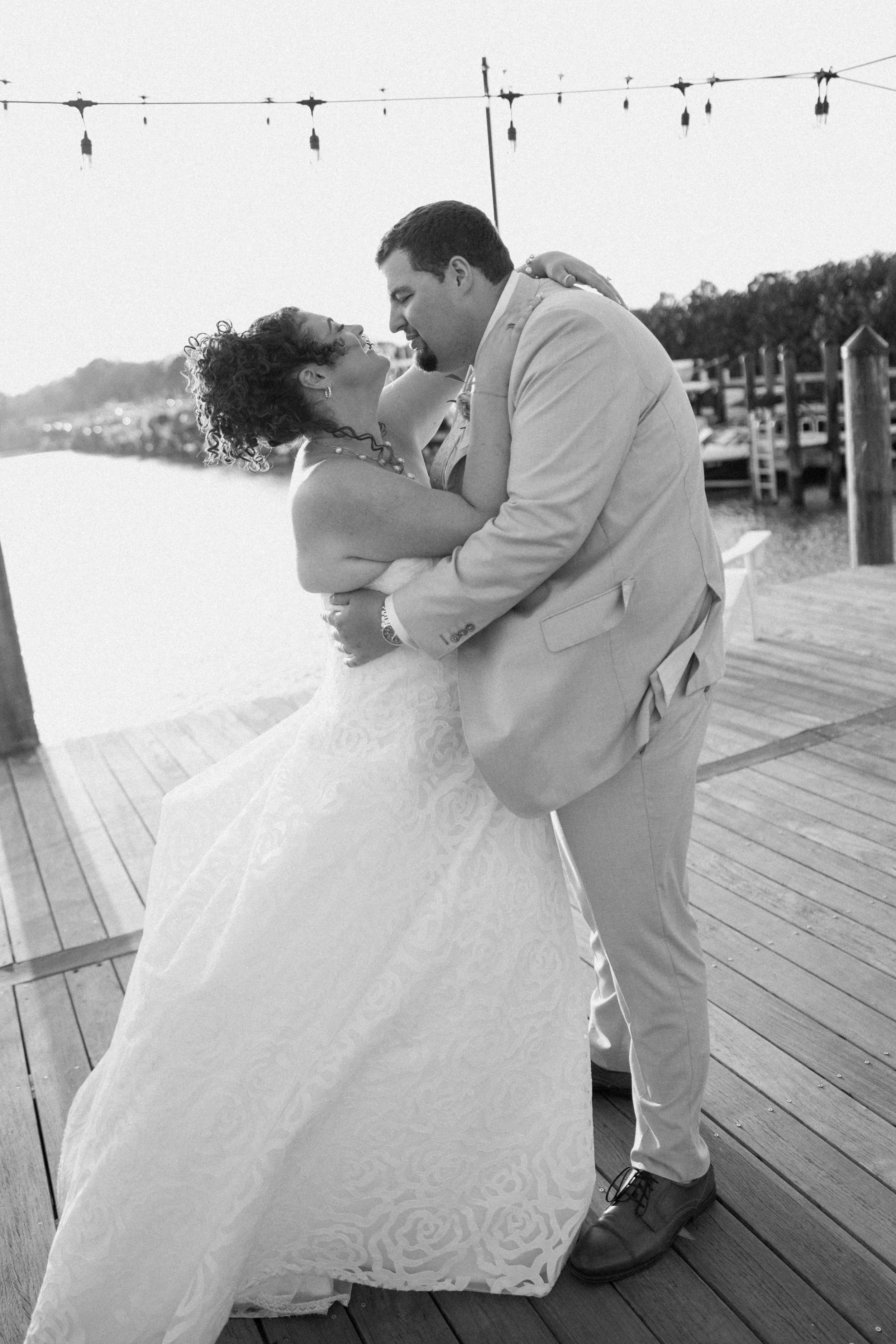 A bride and groom are dancing closely outdoors on a wooden deck, with the groom in a light-colored suit and the bride in a strapless wedding dress, embracing each other affectionately.