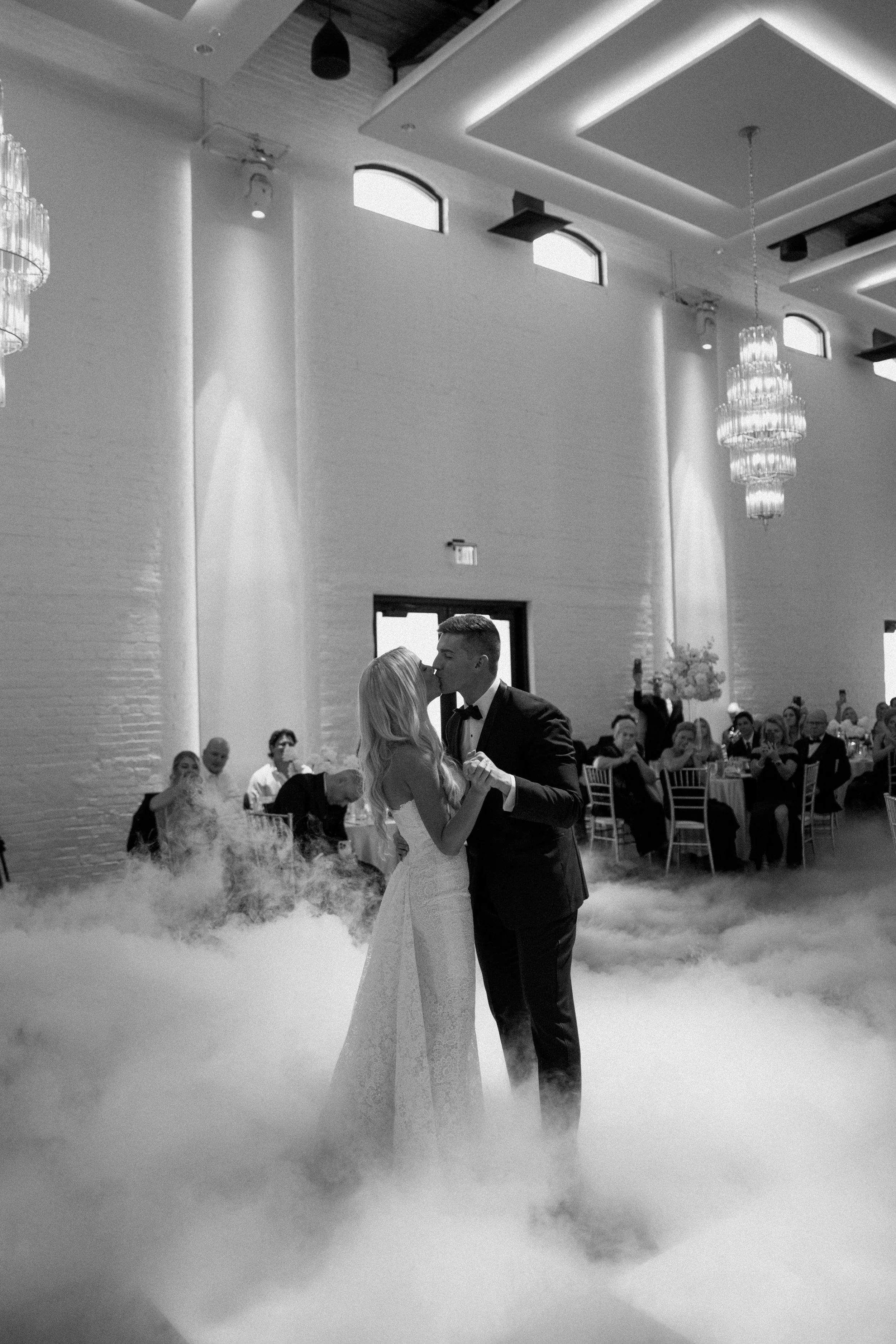 A bride and groom sharing their first dance at their wedding reception in an elegant indoor venue with chandeliers, high windows, and a fog machine.