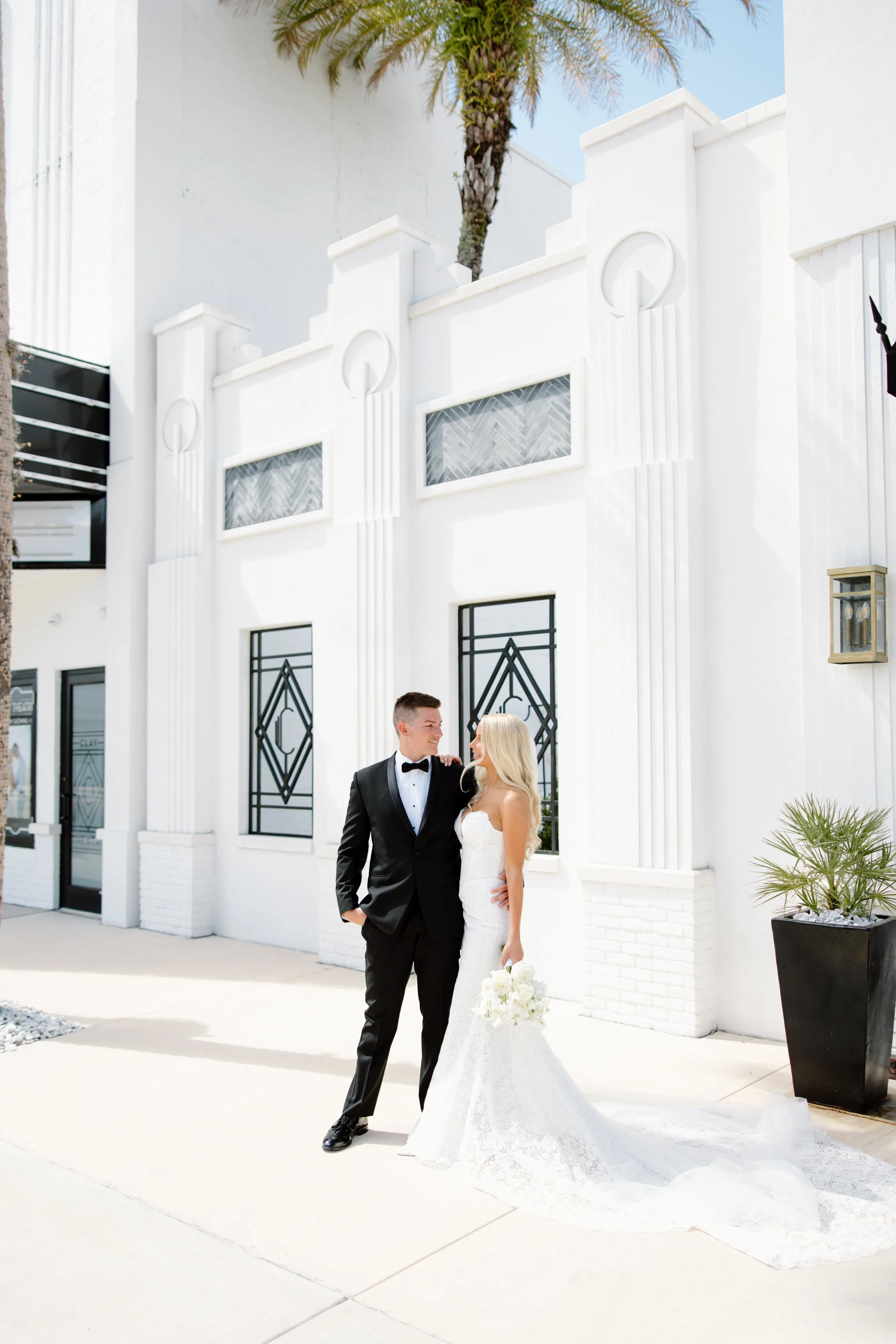 A newlywed couple stands outside a white art deco style building with geometric windows and black doors. The groom is wearing a black tuxedo with a bow tie, and the bride is in a strapless lace wedding gown holding a bouquet. They are smiling at each