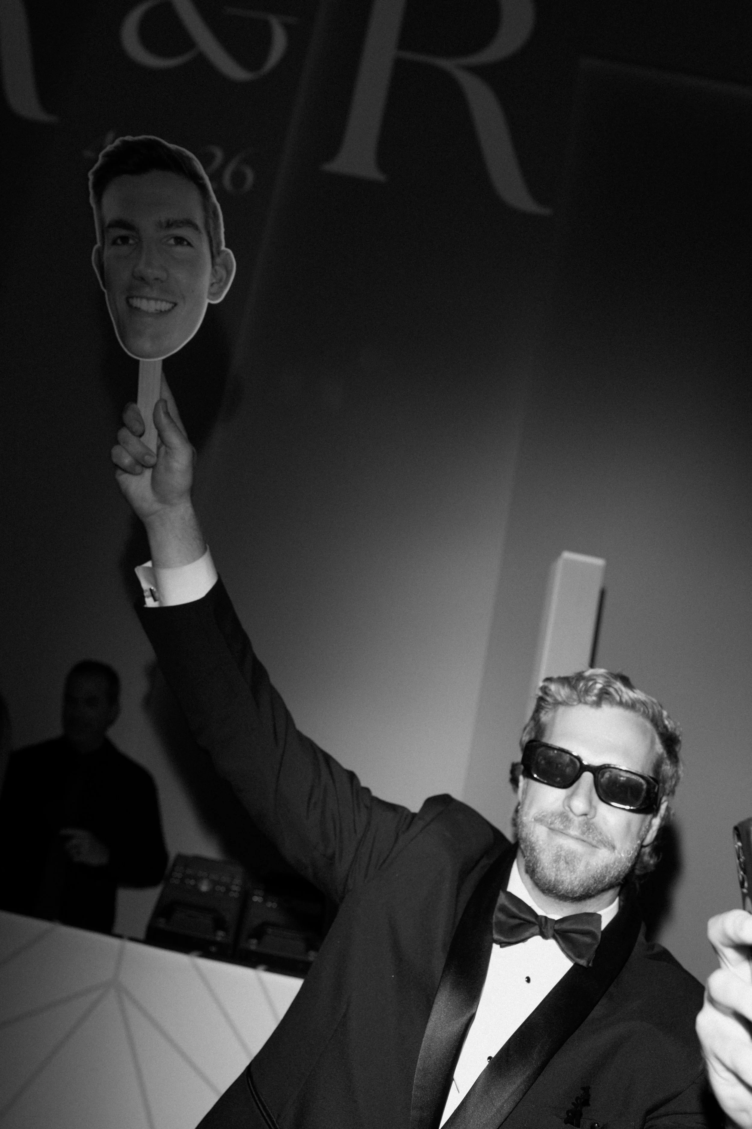 A man in a tuxedo and bow tie, wearing sunglasses, is smiling and raising a cardboard cutout of a person's head with a happy expression.