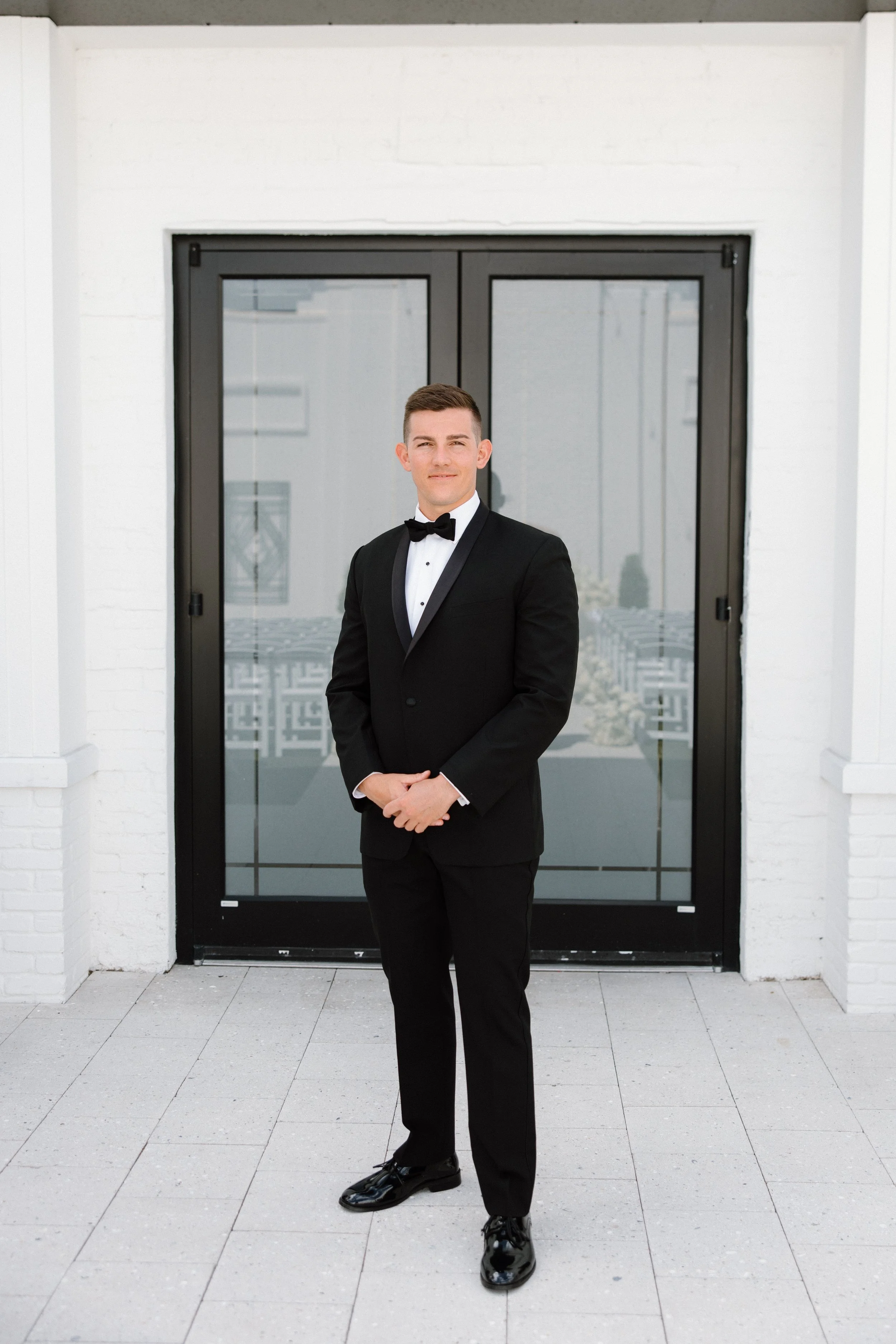 A young man in a black tuxedo with a bow tie standing in front of a modern glass door with a white brick wall surrounding it, his hands clasped in front of him, smiling slightly.