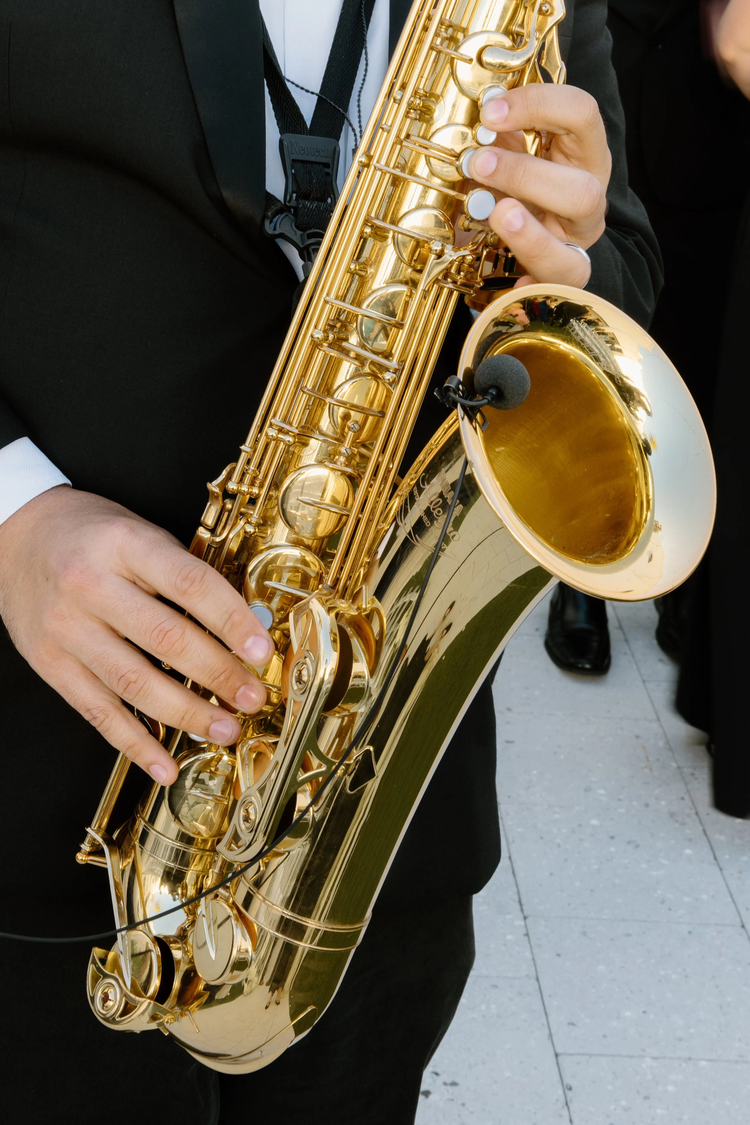 Close-up of a person playing a shiny gold saxophone, with their hands on the instrument and a microphone attached to the bell, wearing a black suit.