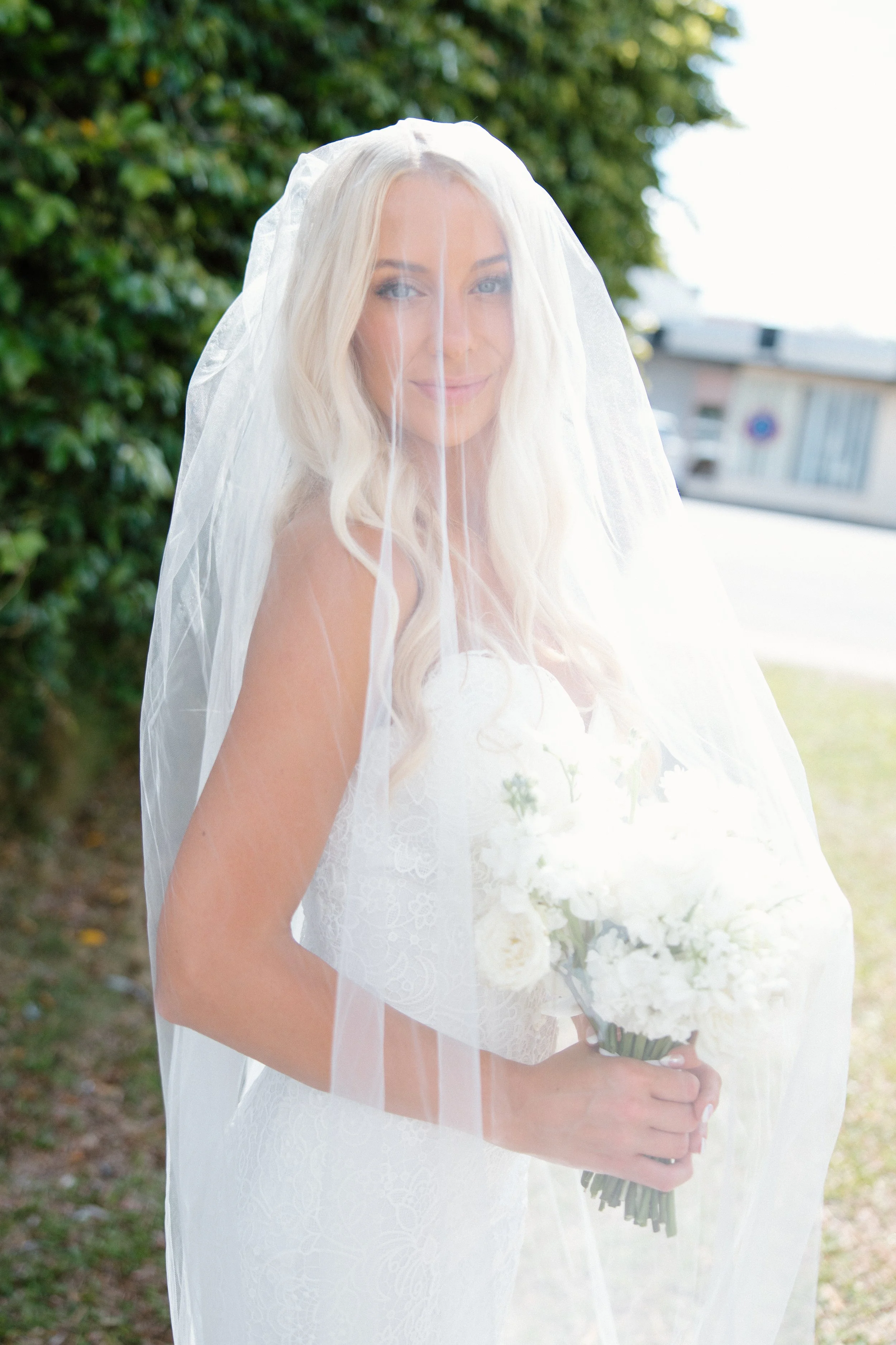 A bride with long blonde hair wearing a white wedding dress and veil, holding a bouquet of white flowers, outdoors with green foliage in the background.