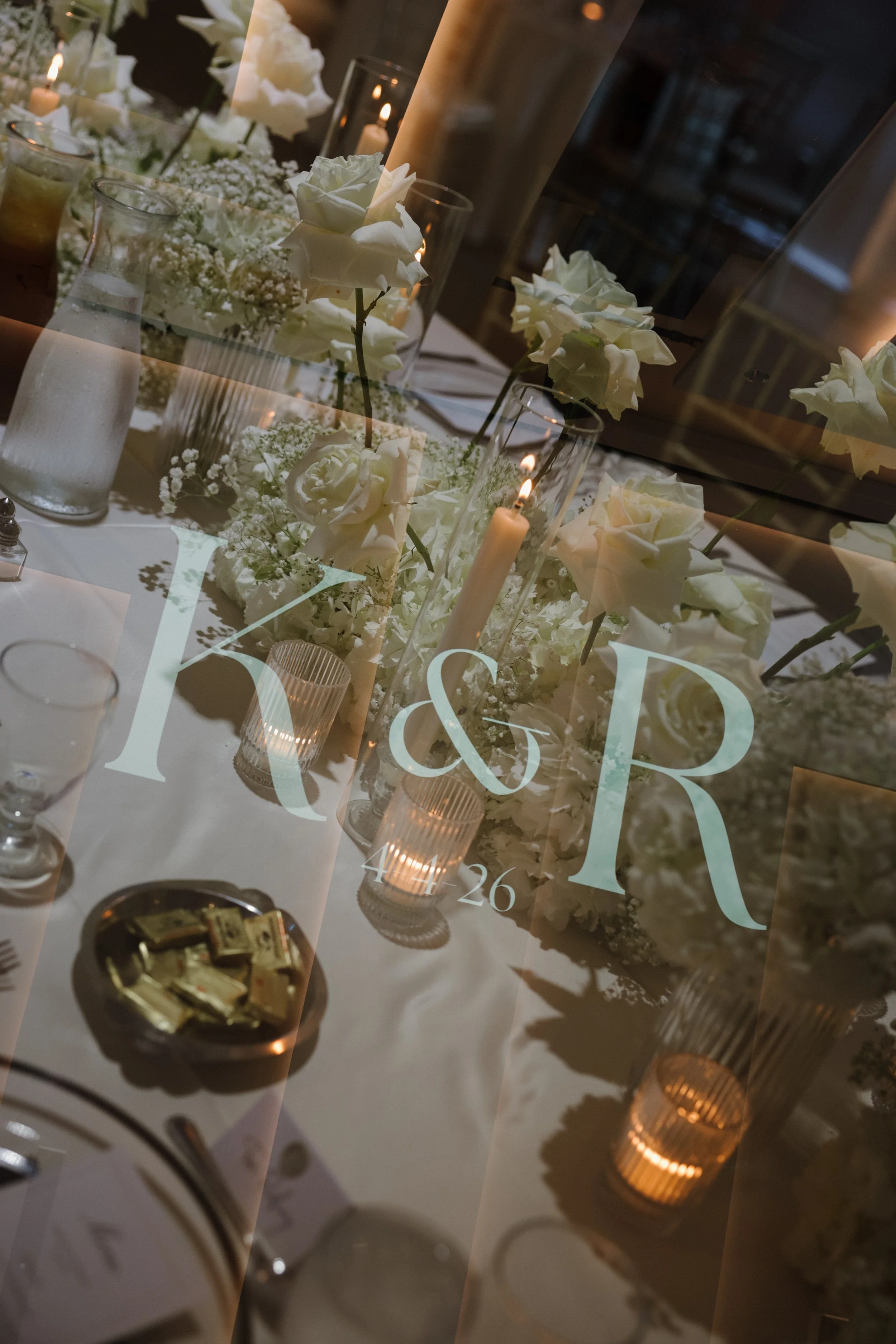 A decorated table with white roses, baby's breath, lit candles, and glassware, visible through a glass with a wedding or event invitation overlay reading 'MR & MRS' and 'April 26'.