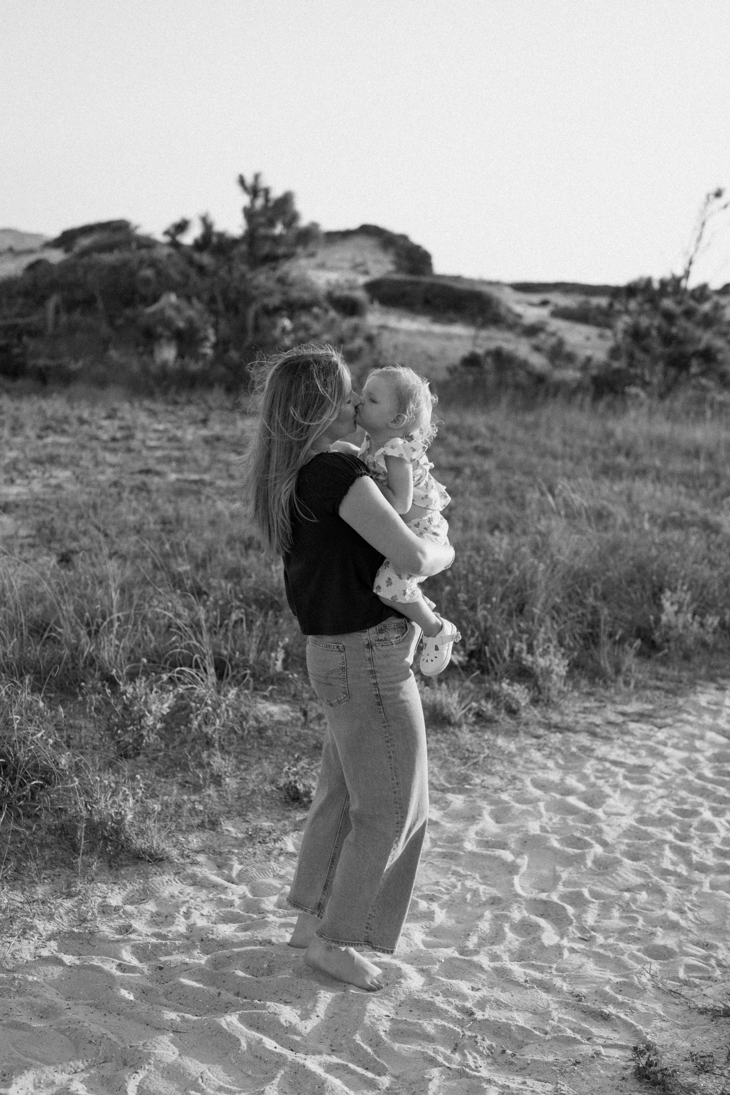 A woman holding a young girl in her arms outdoors on a sandy path, with grassy terrain and rock formations in the background, captured in black and white.