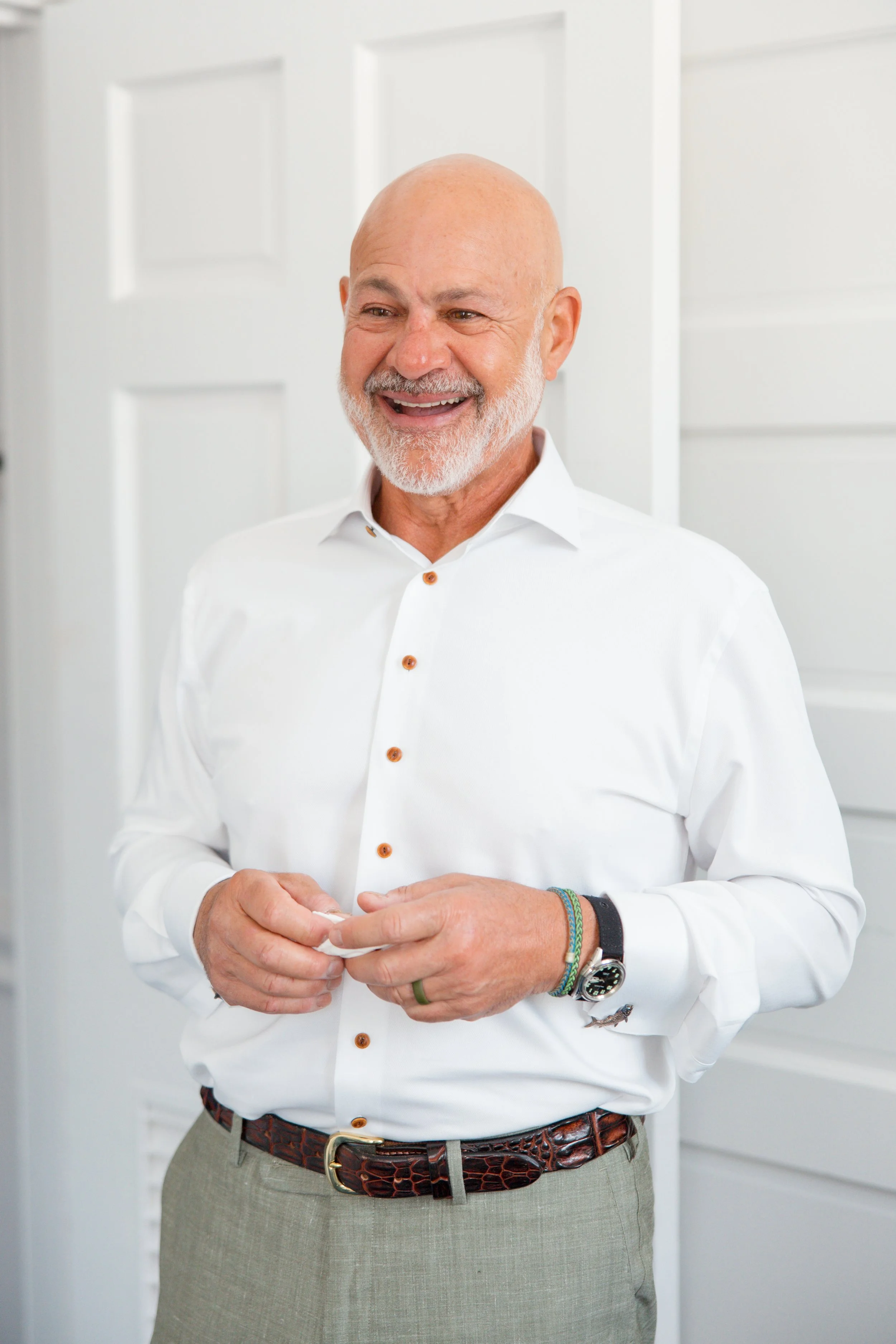 A smiling older man with a beard and a bald head, wearing a white dress shirt with wooden buttons, beige trousers, and a patterned brown belt. He has bracelets on his left wrist and a watch, standing in front of a white background.