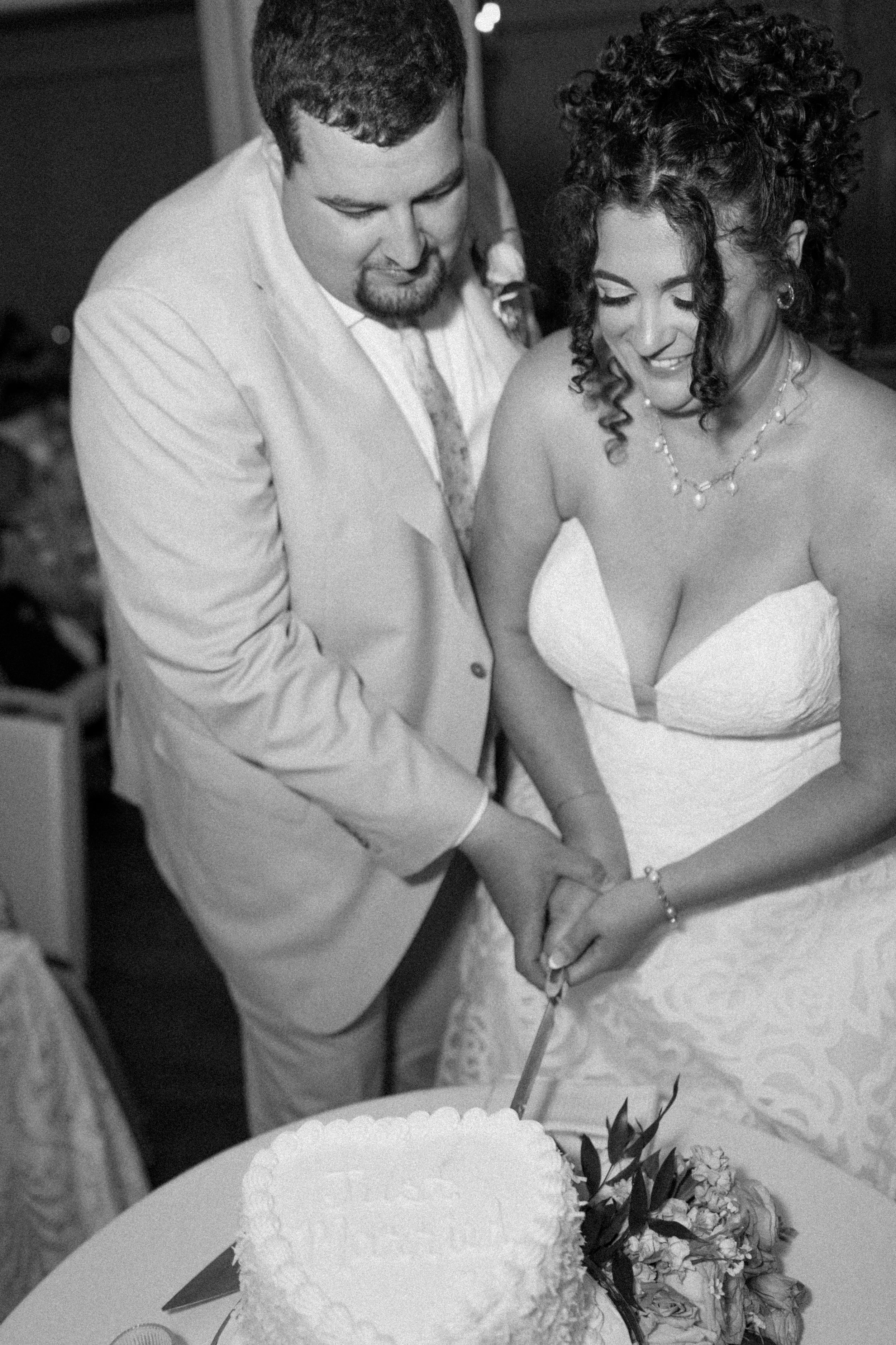 A couple cutting a wedding cake together at their wedding reception, with a decorated cake and floral arrangements on the table.