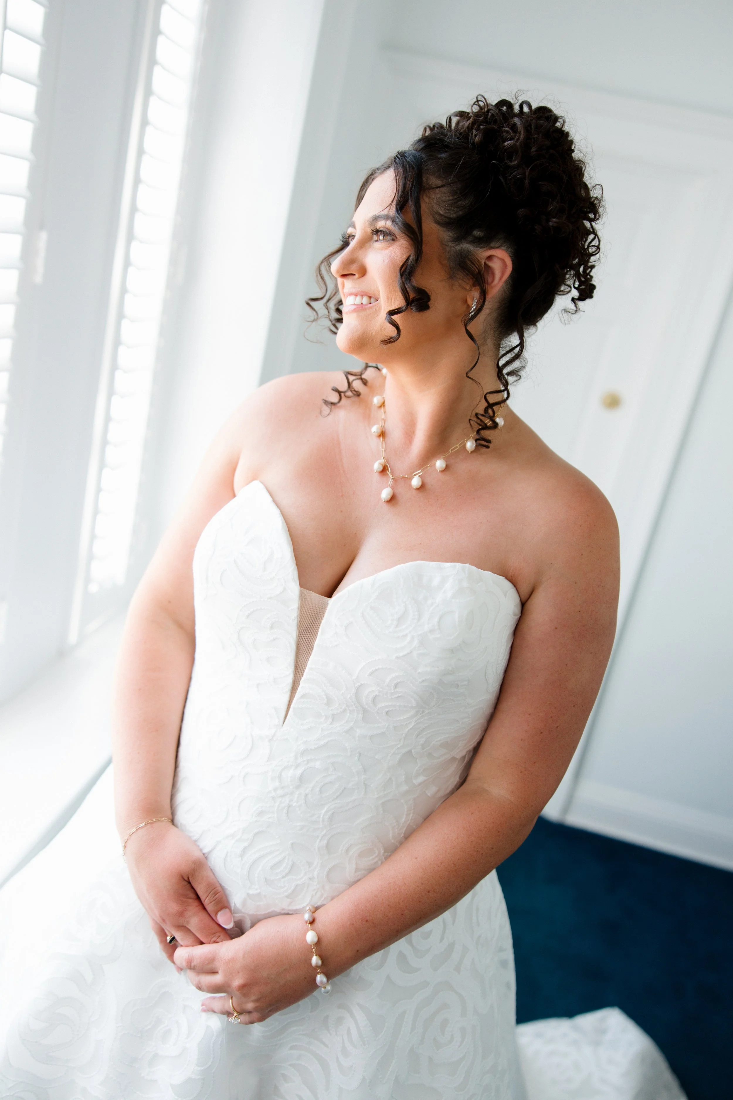 A woman in a white wedding dress with curly dark hair, wearing pearl jewelry, standing indoors by a window with shutters, smiling and looking to the side.