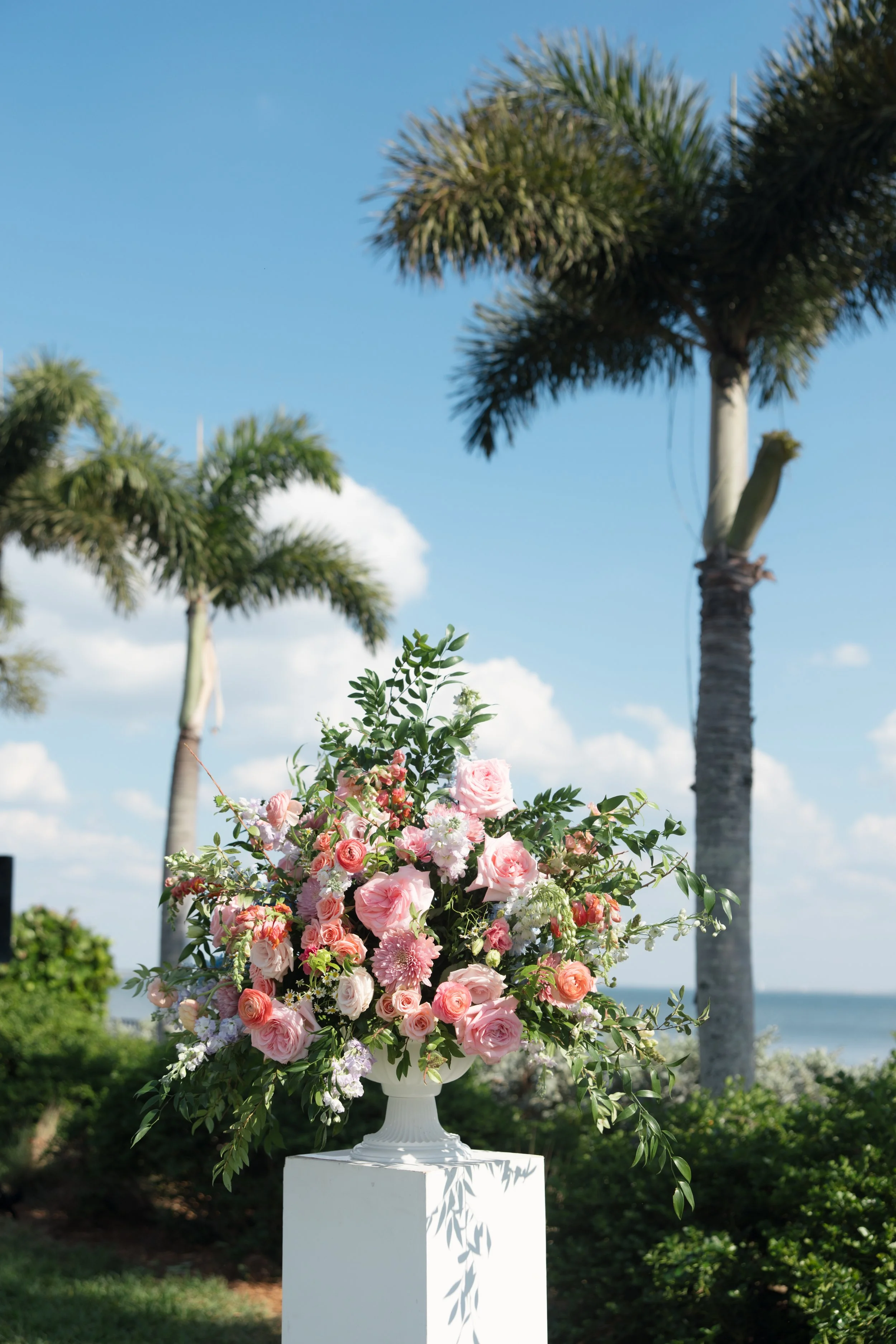 Flower arrangement with pink and white roses, placed on a white pedestal outdoors with palm trees and blue sky in the background.