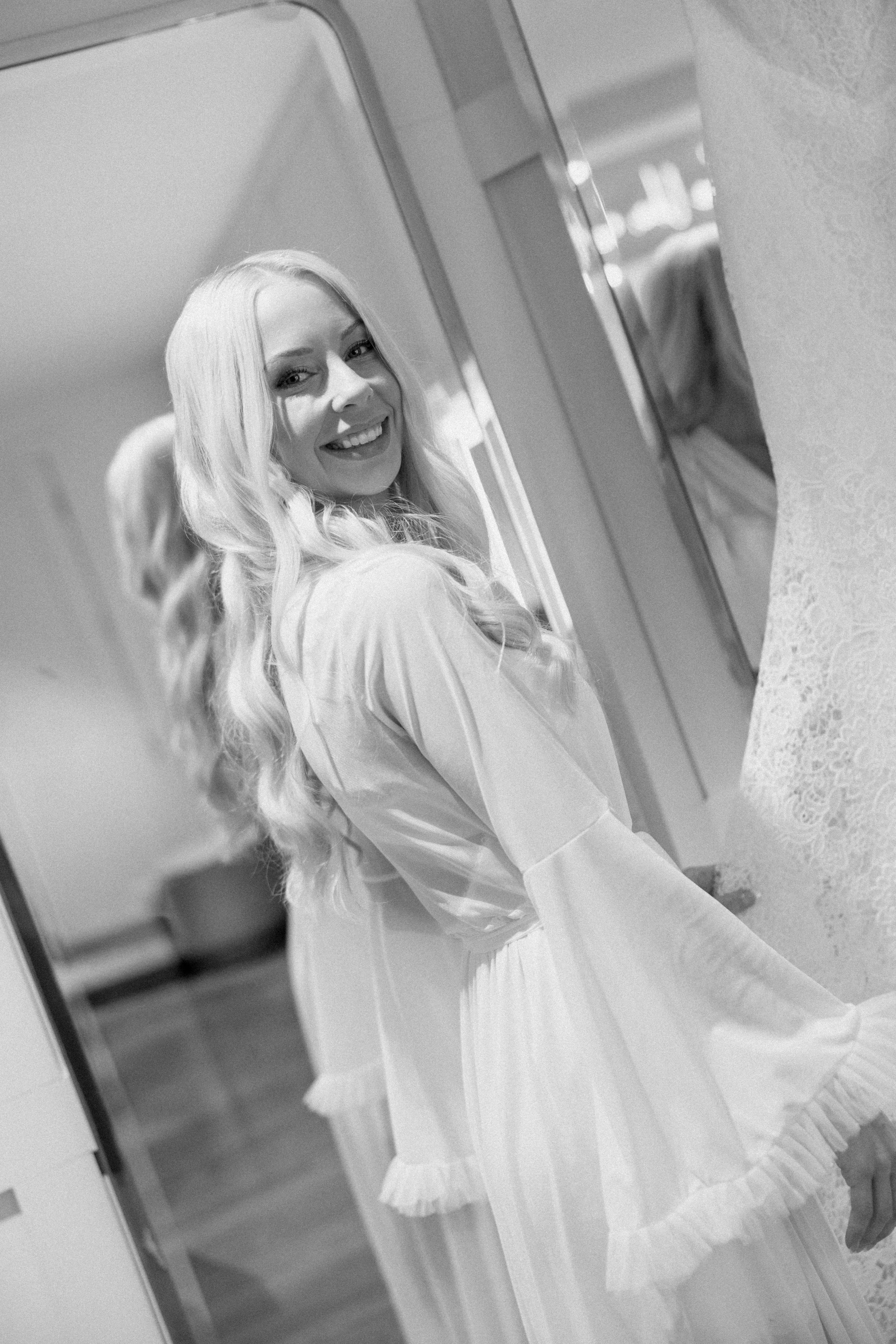 A woman with long blonde hair smiling, dressed in a light-colored dress with lace details, standing in front of a mirror in a softly lit room.