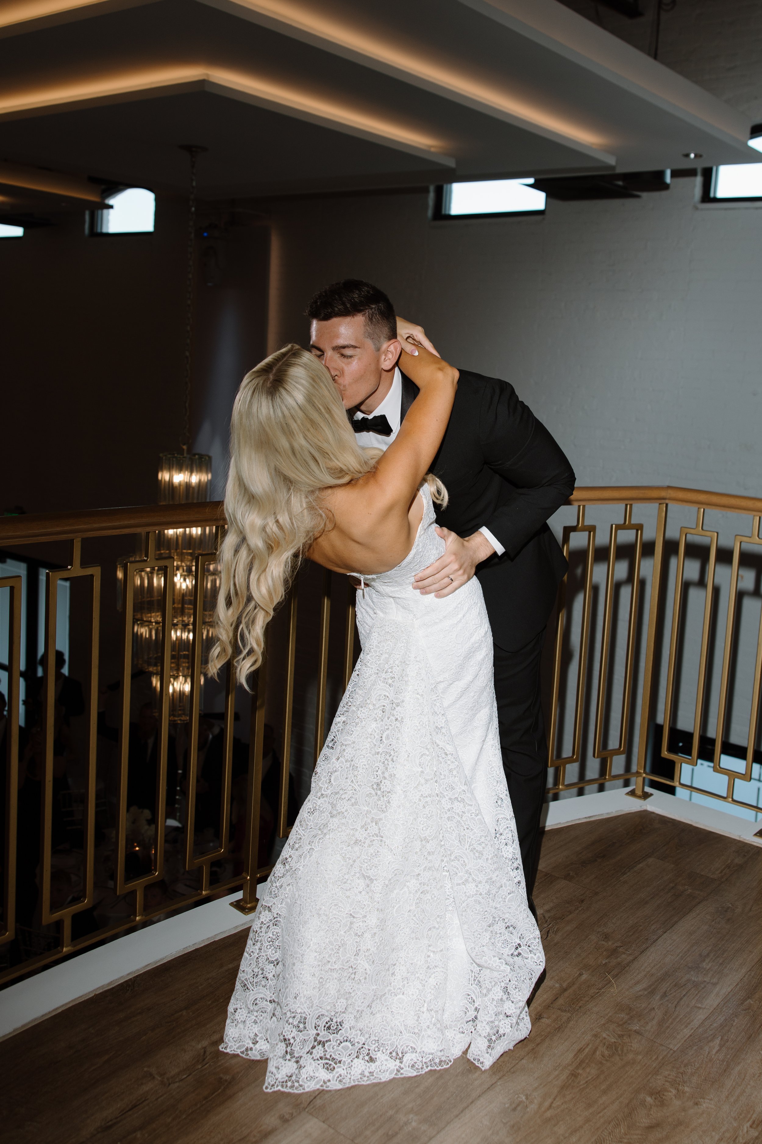 A newlywed couple sharing a kiss on a staircase at a wedding reception, with the groom in a black tuxedo and the bride in a white lace wedding dress, in an indoor venue with wood flooring and a decorative gold railing.
