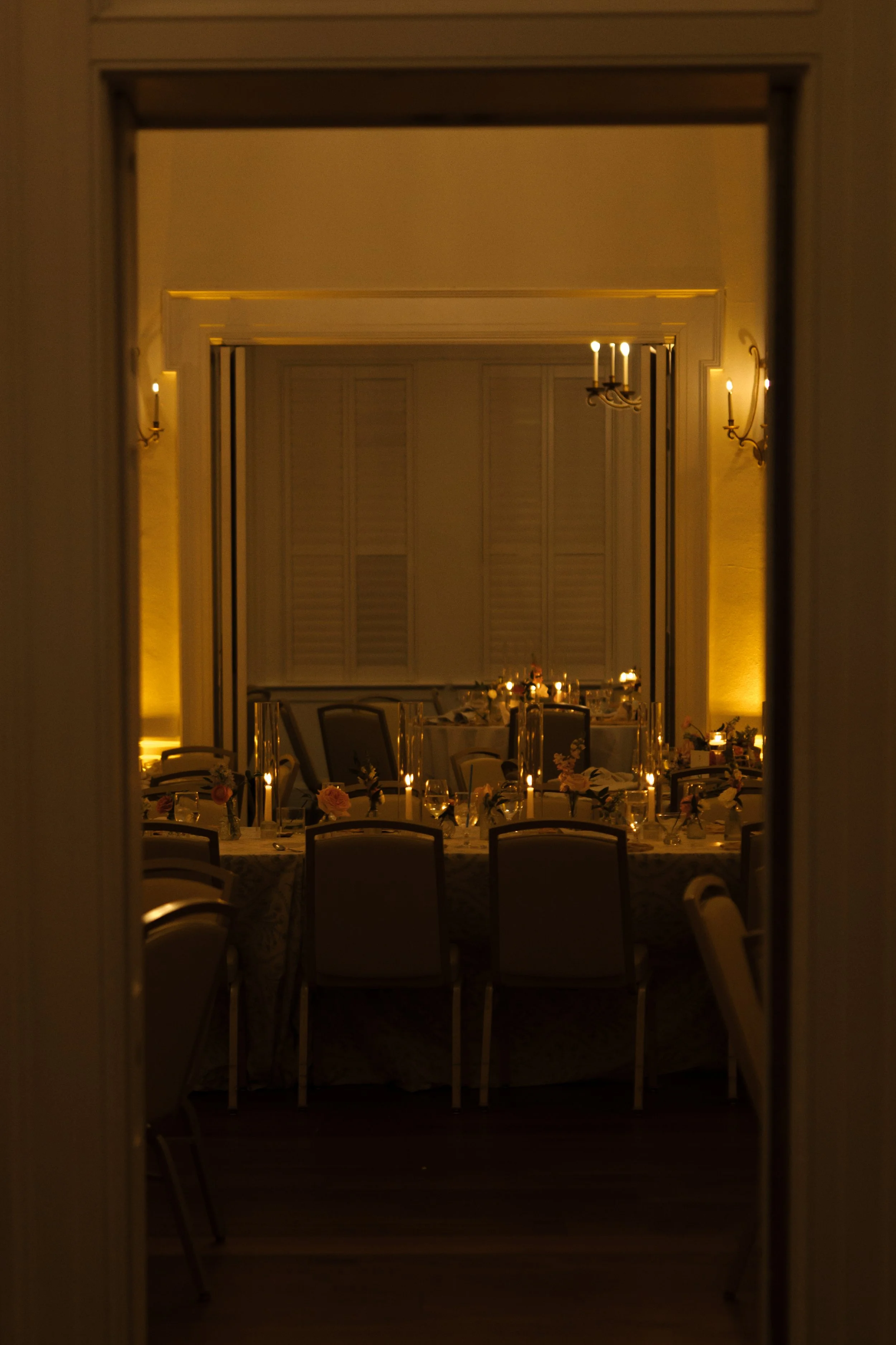 A dining room with elegant chandelier lighting, decorated for a formal event, with round tables set with floral centerpieces, candles, and glassware, viewed through a doorway.