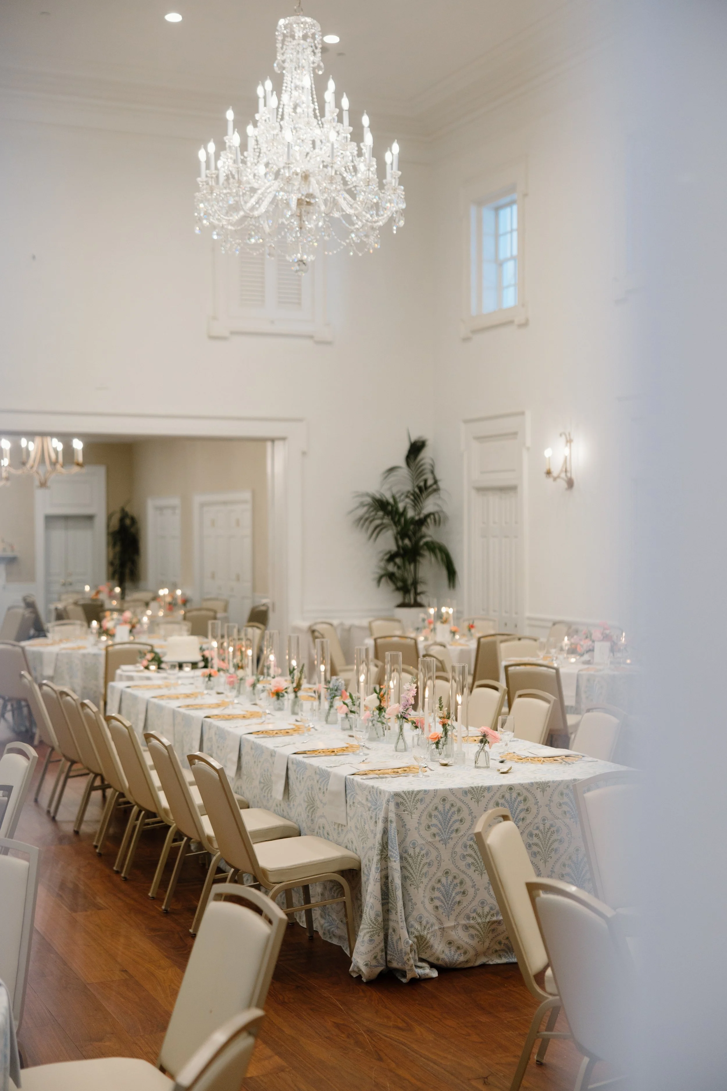 Elegant banquet hall decorated for a celebration with a chandelier, round tables with floral centerpieces, and chairs arranged around the tables.