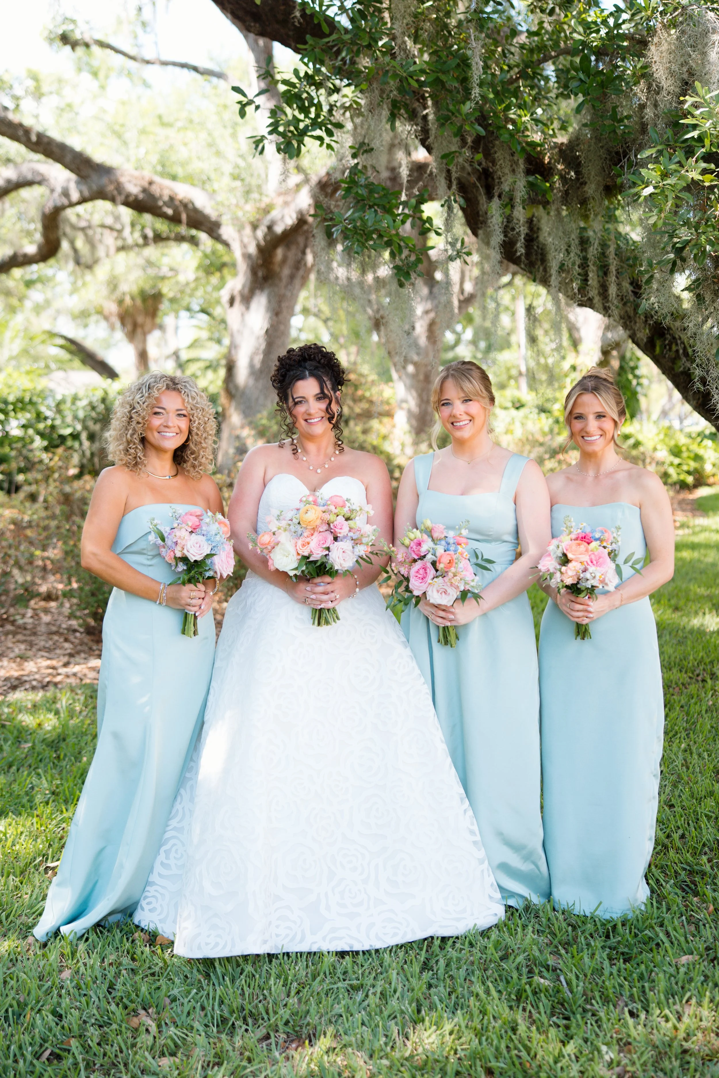 Four women dressed as bridesmaids in light blue dresses holding flower bouquets, standing outdoors under a large tree with green leaves and Spanish moss, during daytime.