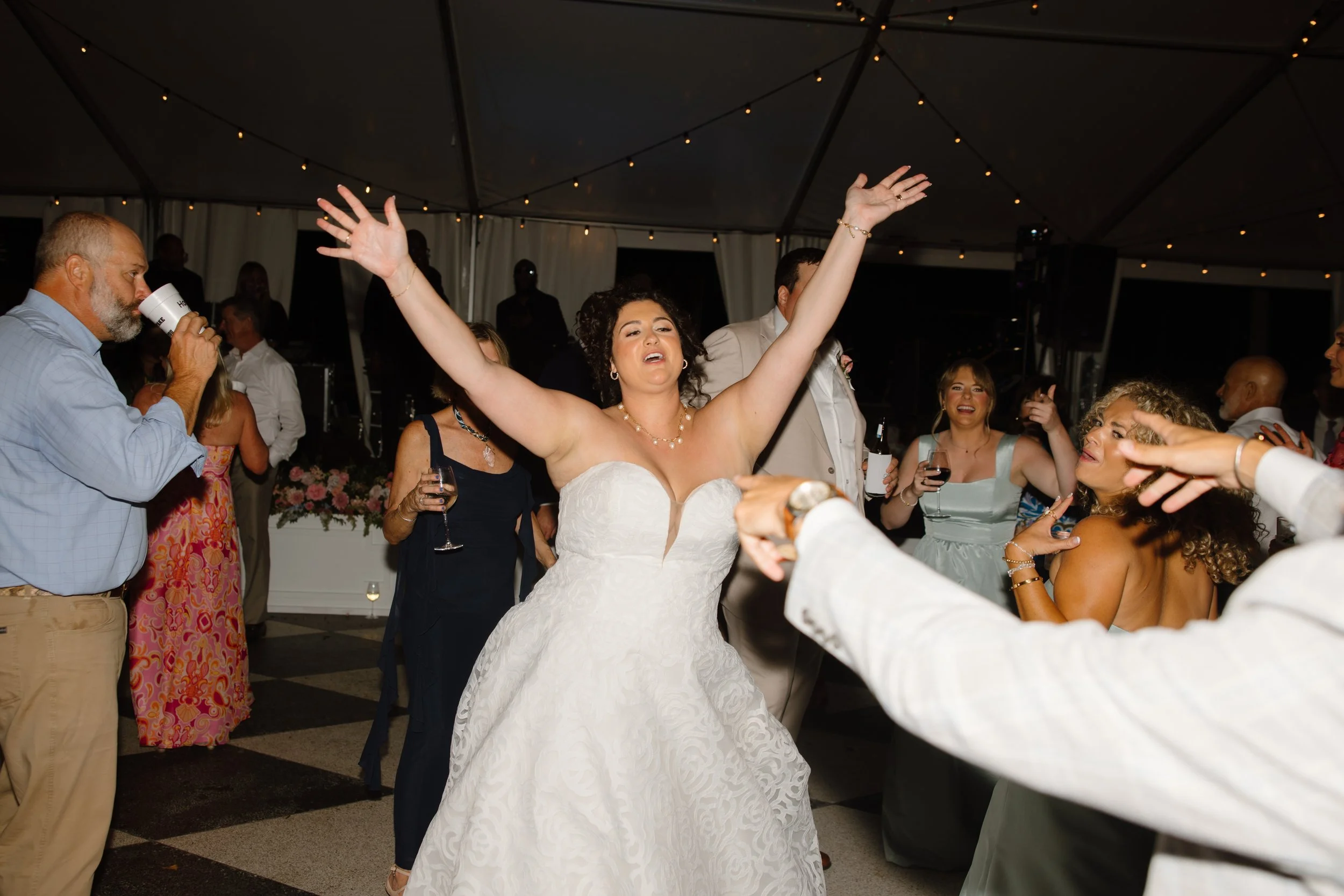 A bride dancing with her arms raised at her wedding reception, surrounded by guests who are drinking and smiling inside a decorated venue with string lights.