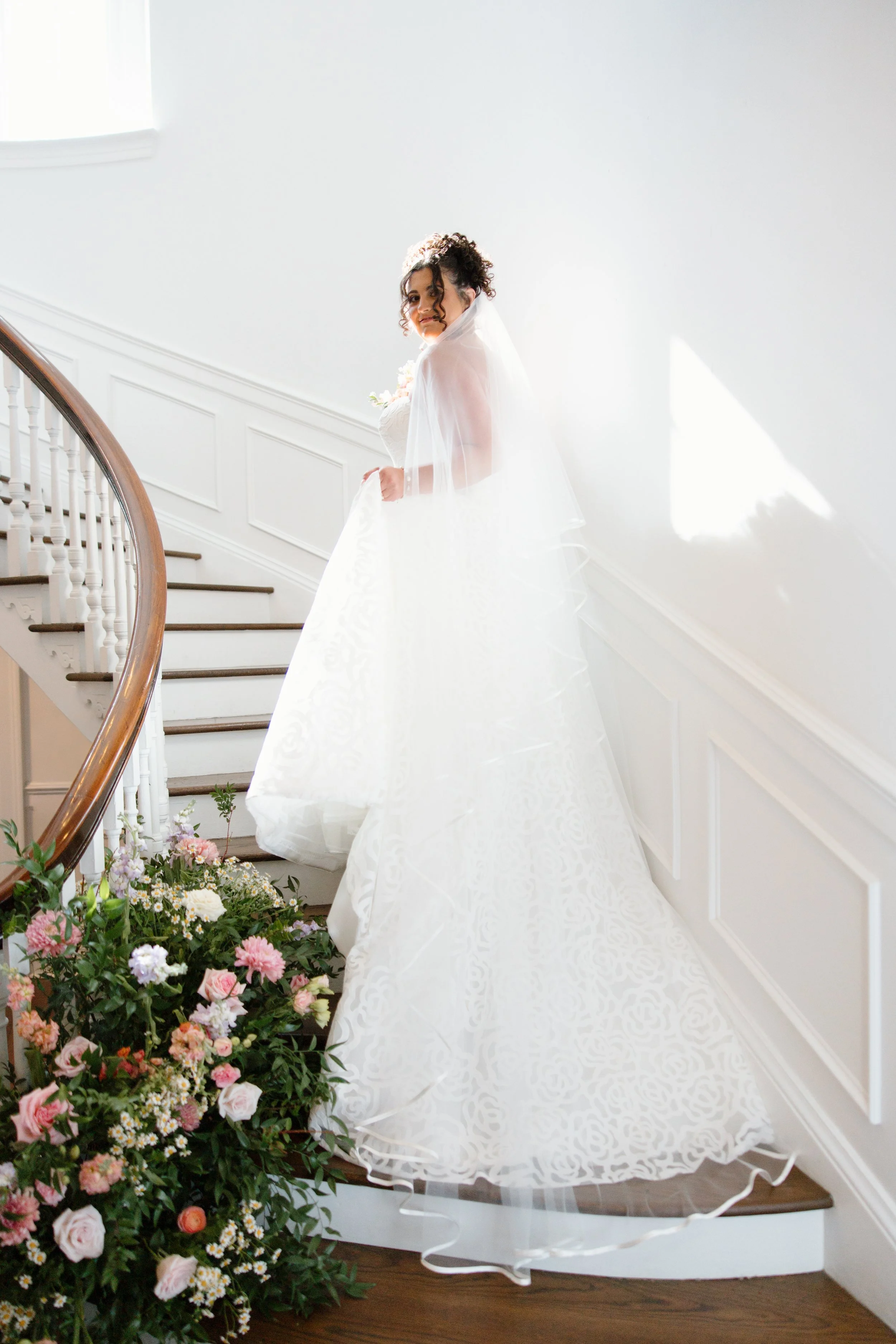 A bride wearing a white wedding gown and veil standing on a staircase, surrounded by colorful flowers.