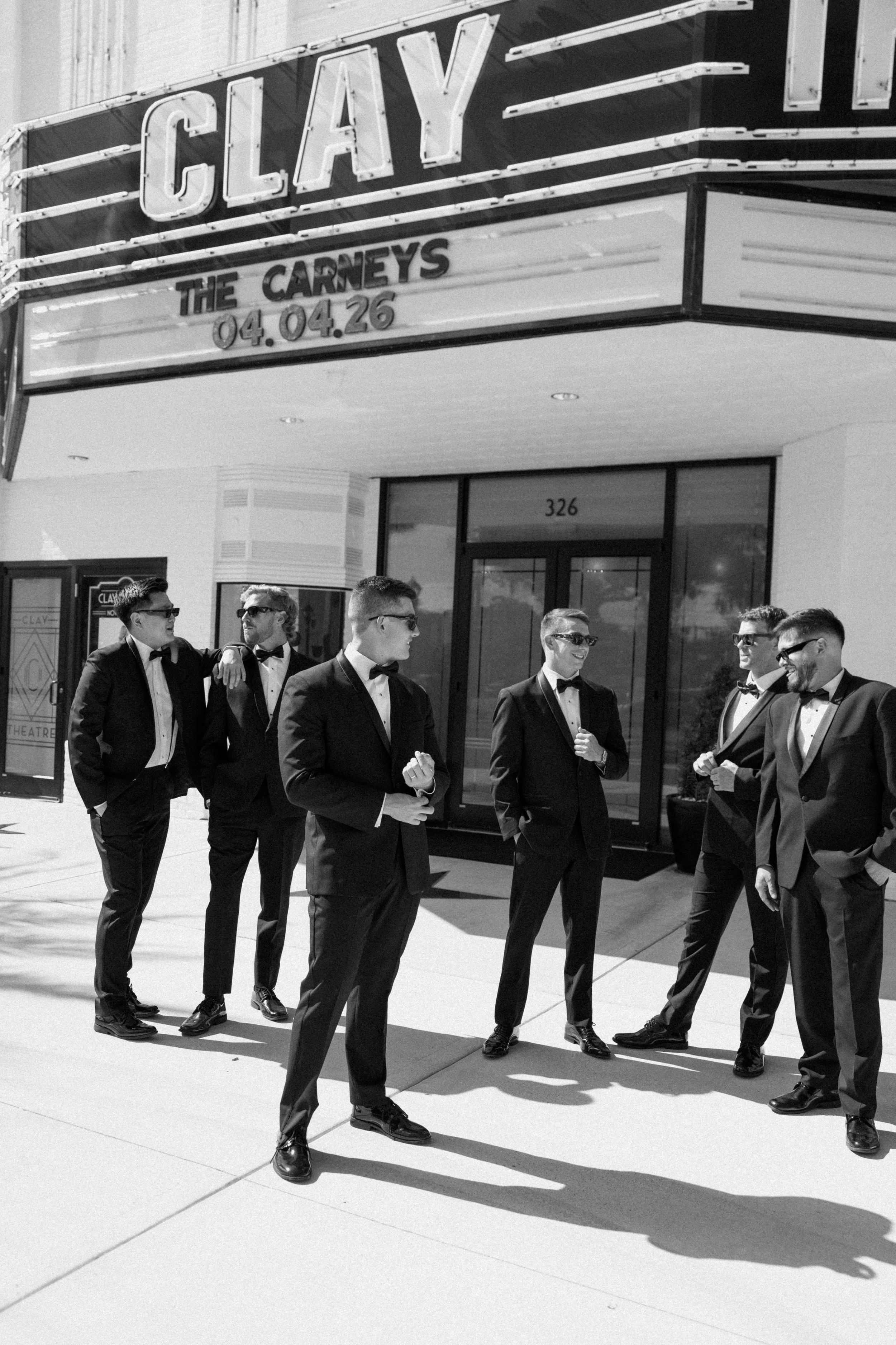A group of men in tuxedos and sunglasses walking outside a theater with a marquee advertising a show titled "The Carneys" on April 26, 2026.