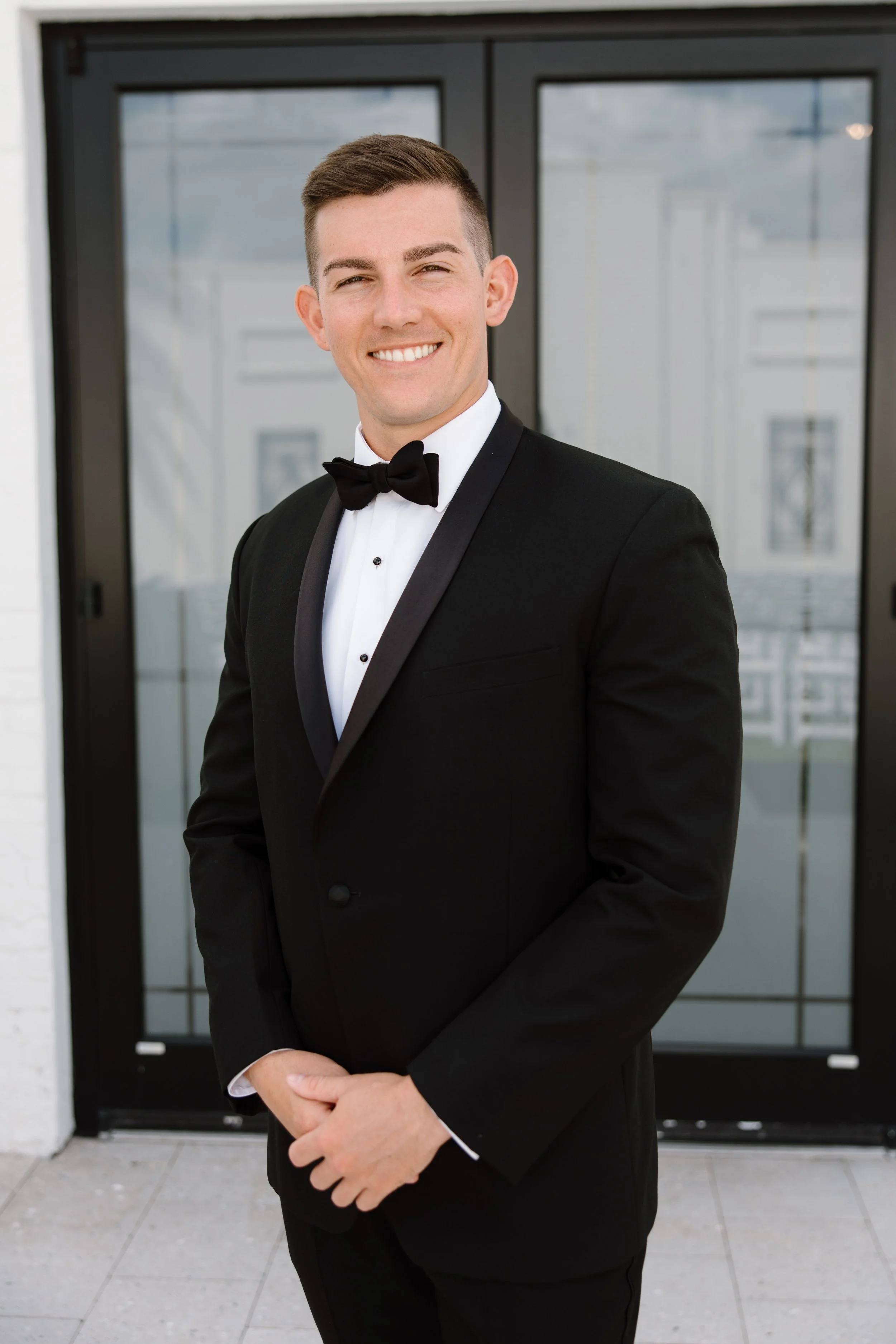 A young man in a tuxedo with a bow tie standing outside in front of glass doors, smiling with arms crossed.