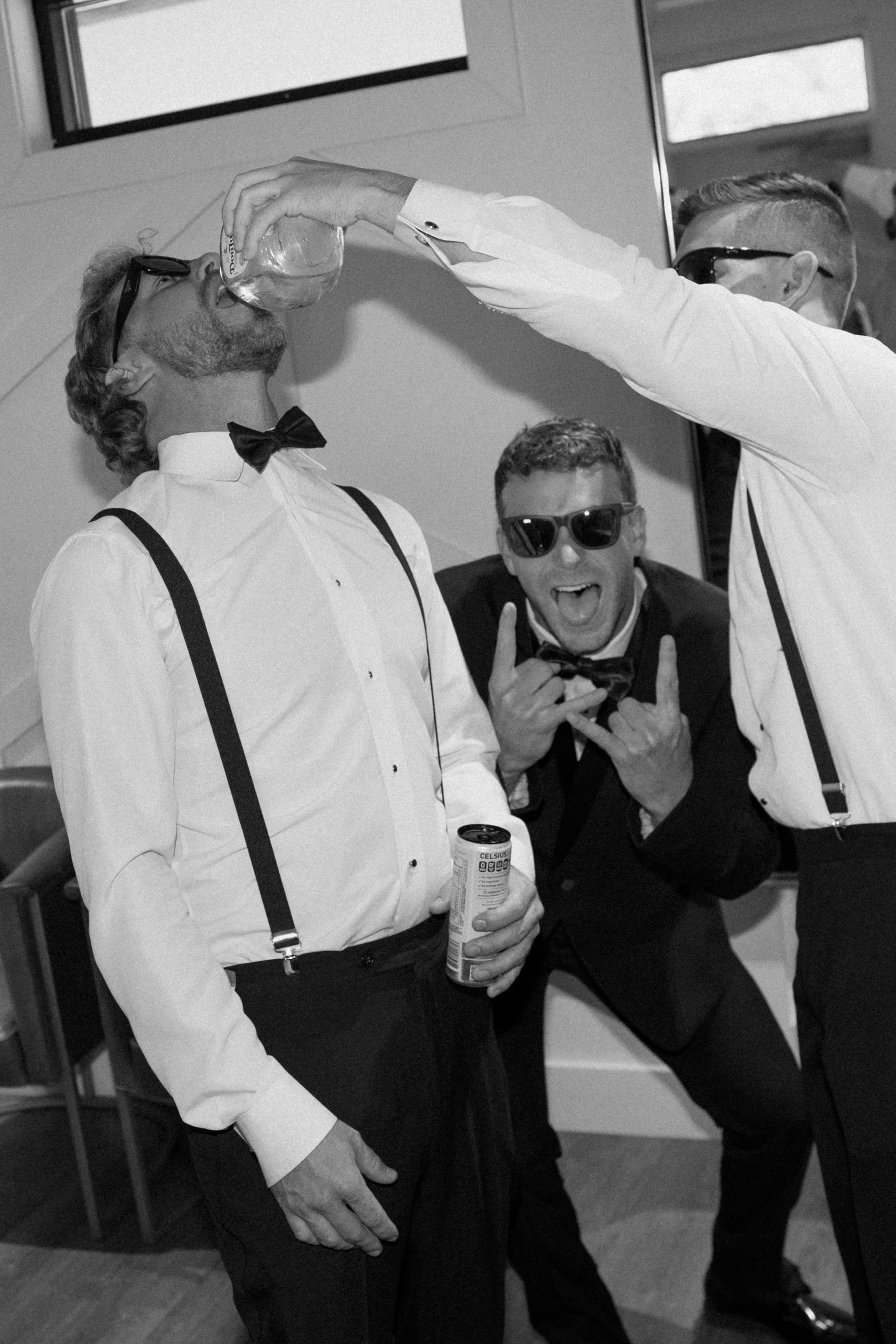 Three men dressed in tuxedos and bow ties, two of them wearing sunglasses, are at a celebration or party. One man is holding a tissue can and is drinking from a glass, while the other is extending his arm to pour a drink into the first man's mouth. T