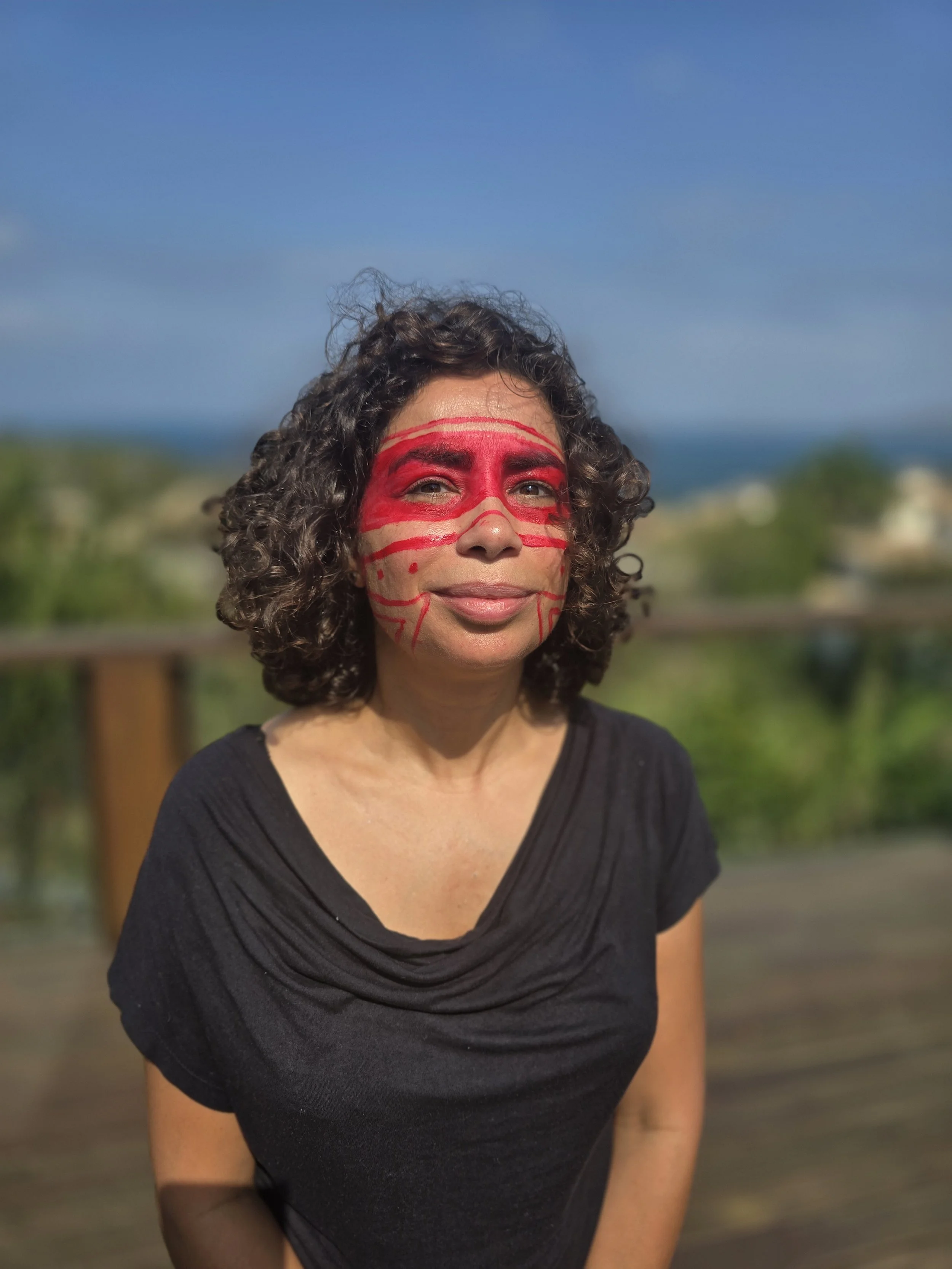 A woman with curly hair and a black shirt smiling outdoors. Her face is painted with red lines and dots resembling a mask.
