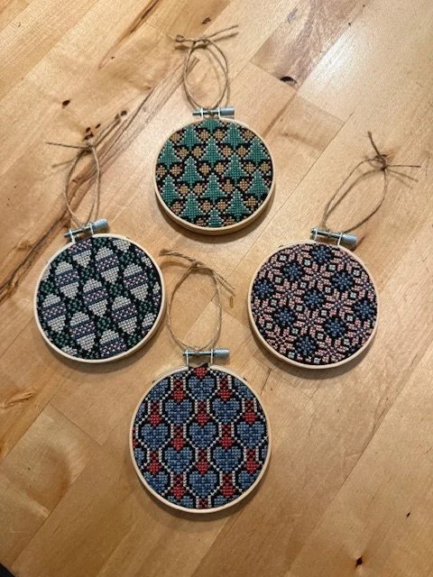 Four round embroidery hoops with cross-stitch patterns in various colors, laid out on a wooden surface. Each hoop features a different intricate geometric design.